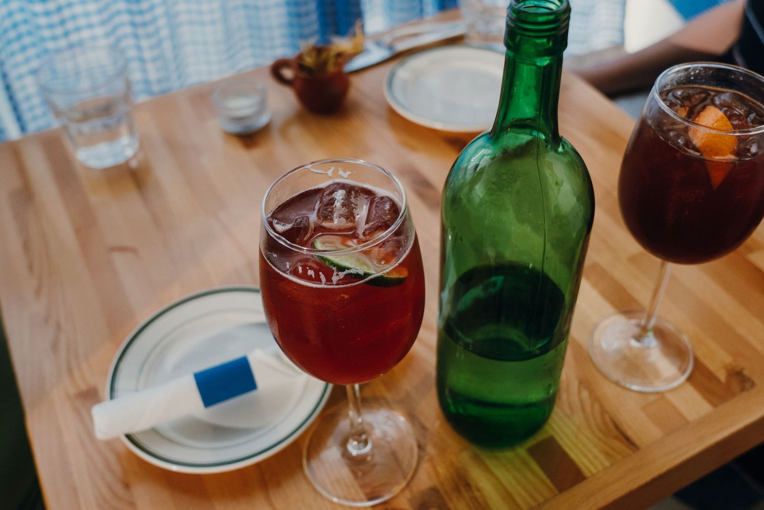 Red wine spritz cocktail in a large glass next to a green bottle on a wooden restaurant table.