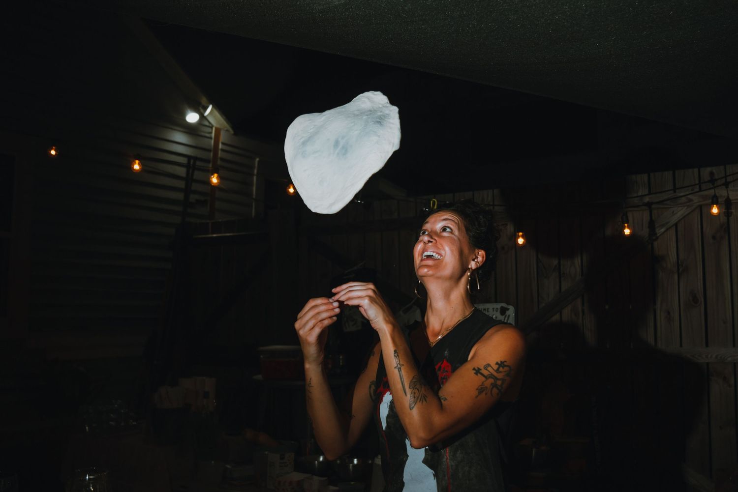 Tattooed woman tossing pizza dough in the air under string lights at an outdoor event.