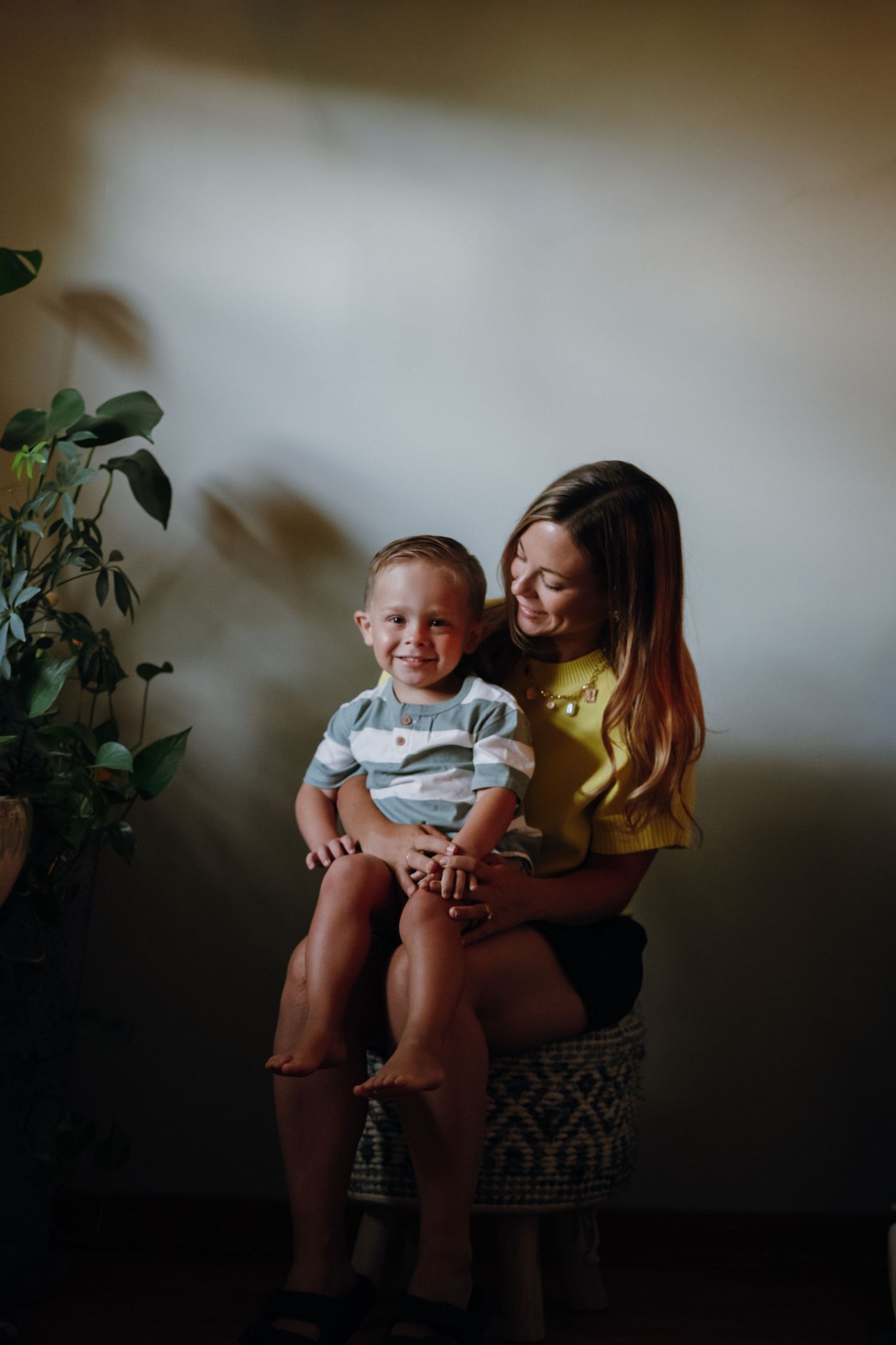 Smiling toddler boy sitting on mother's lap in a warmly lit indoor portrait session.