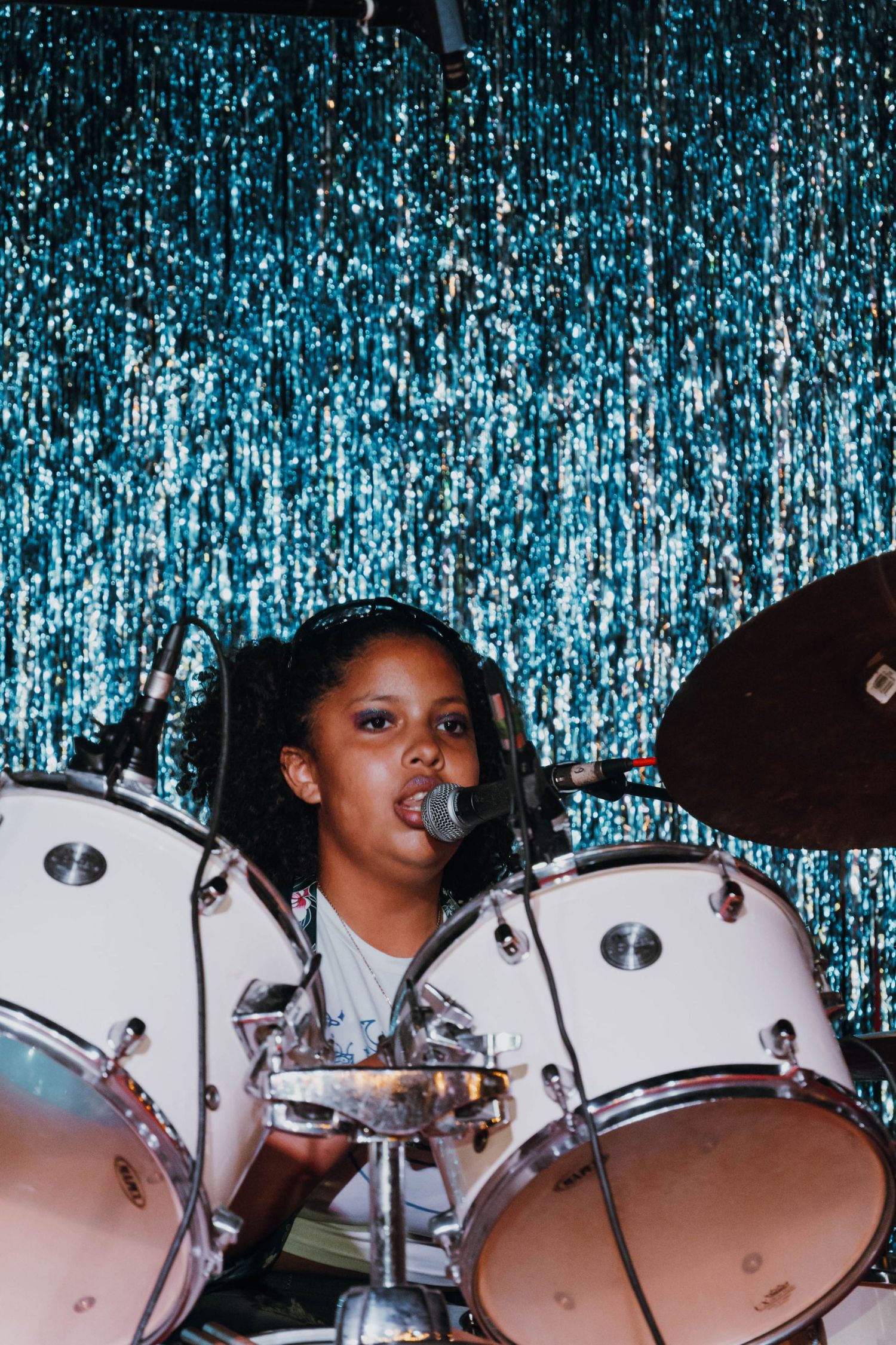 Young girl singing and playing drums on stage with a glittery blue curtain backdrop.