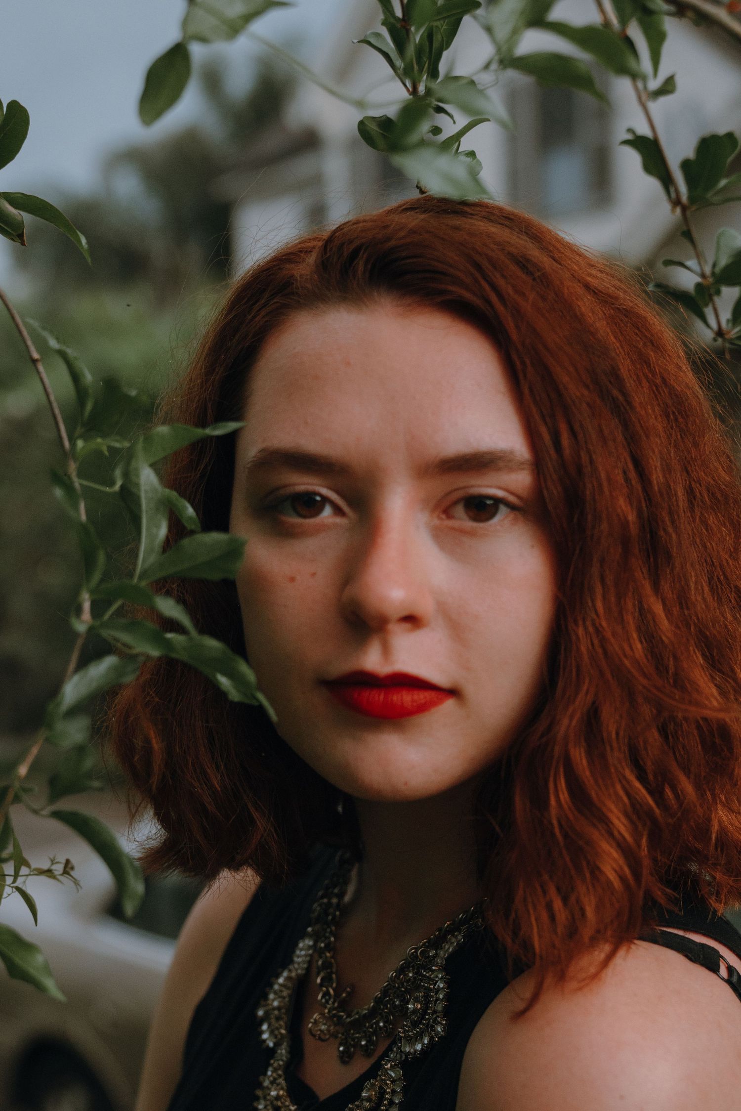 Close portrait of young woman with auburn hair and red lips framed by green leaves outdoors.