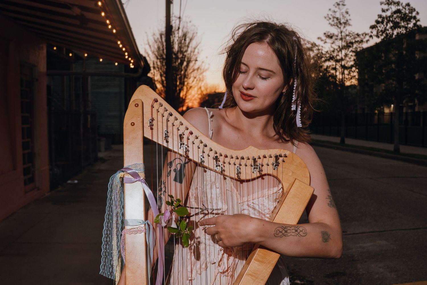 Young woman with tattoos plays a wooden harp at sunset on a city street with string lights.