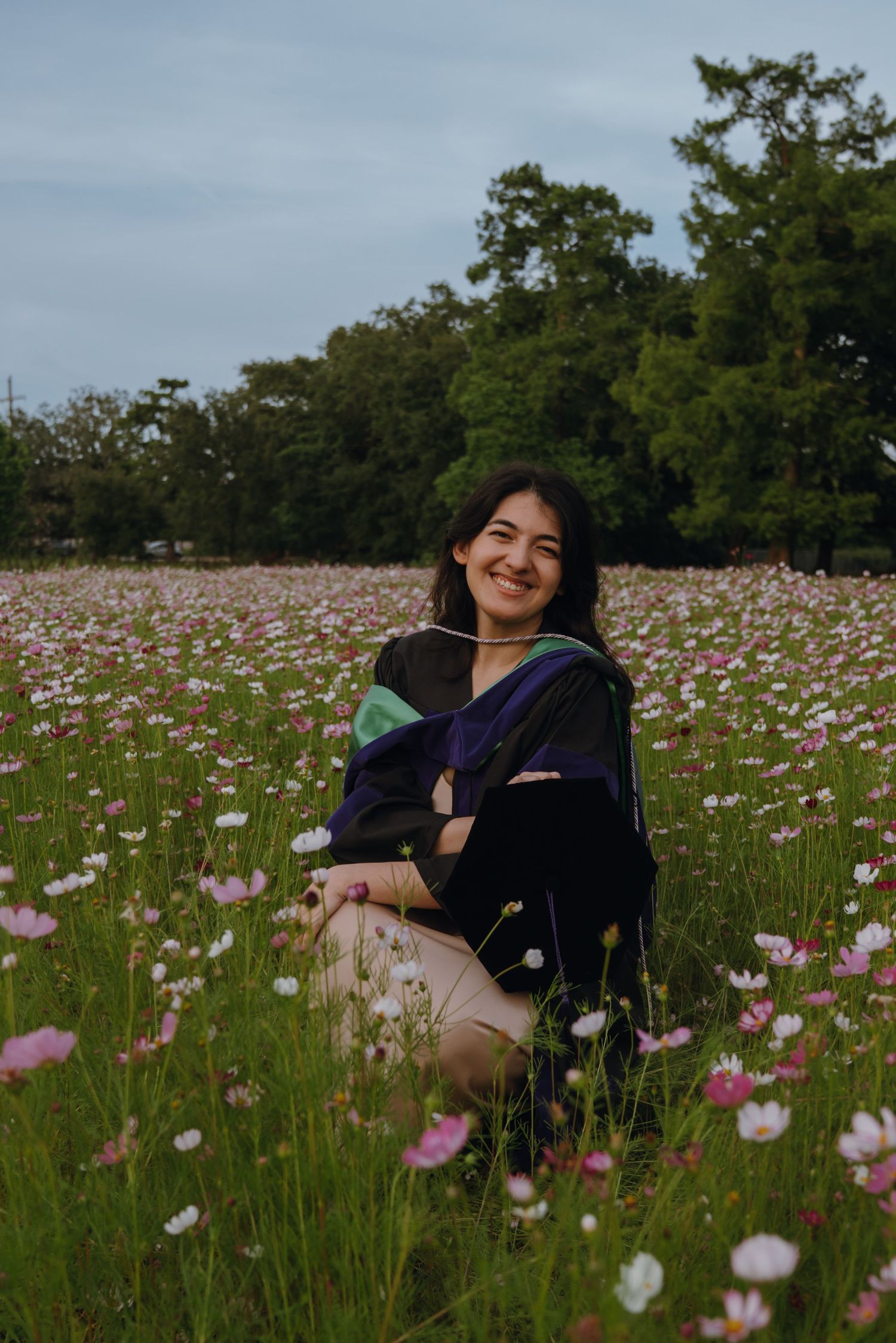Smiling graduate in academic regalia sitting among pink and white cosmos flowers in field.