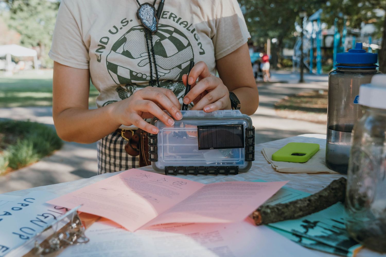 Woman in graphic tee operating a portable emergency radio at an outdoor table with open notebooks.