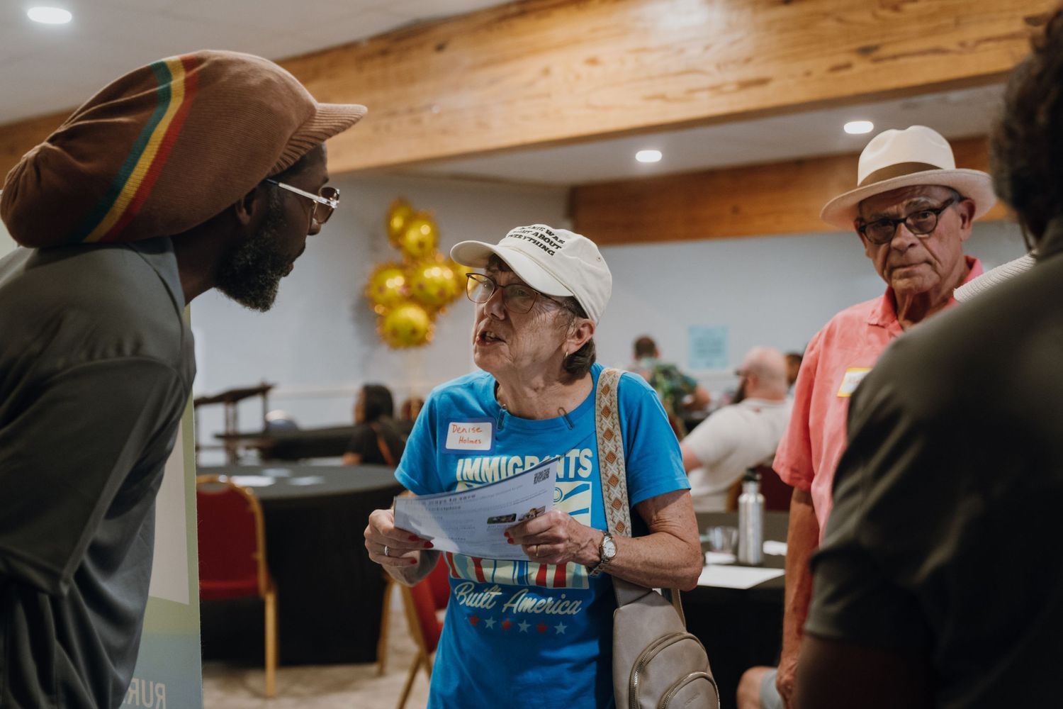 People engaged in conversation at an indoor community gathering with gold balloon decorations.