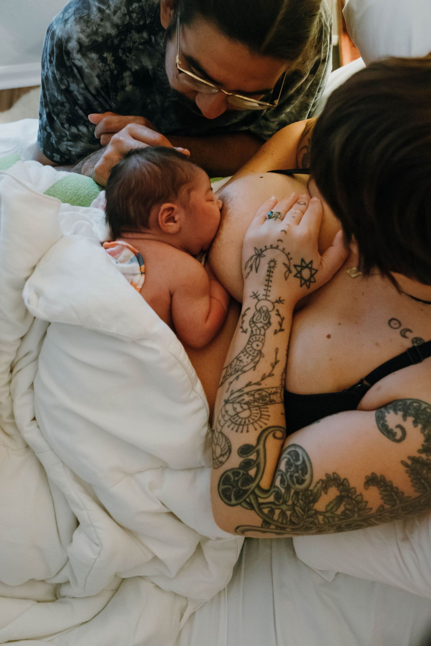 Tattooed mother breastfeeding newborn baby in bed assisted by midwife after home birth.