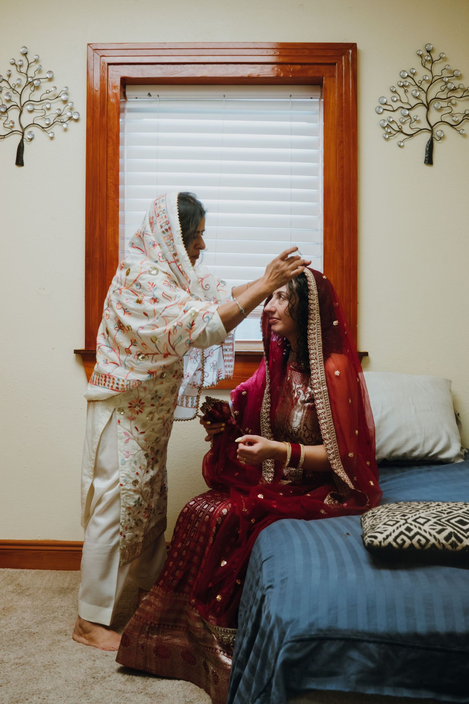 Elder woman adjusts red dupatta on seated South Asian bride in traditional red bridal lehenga.