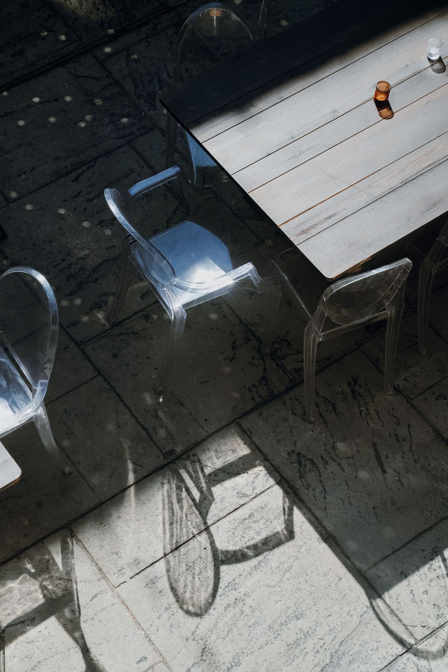 Aerial view of acrylic chairs casting shadows beside a wooden table on stone pavement.
