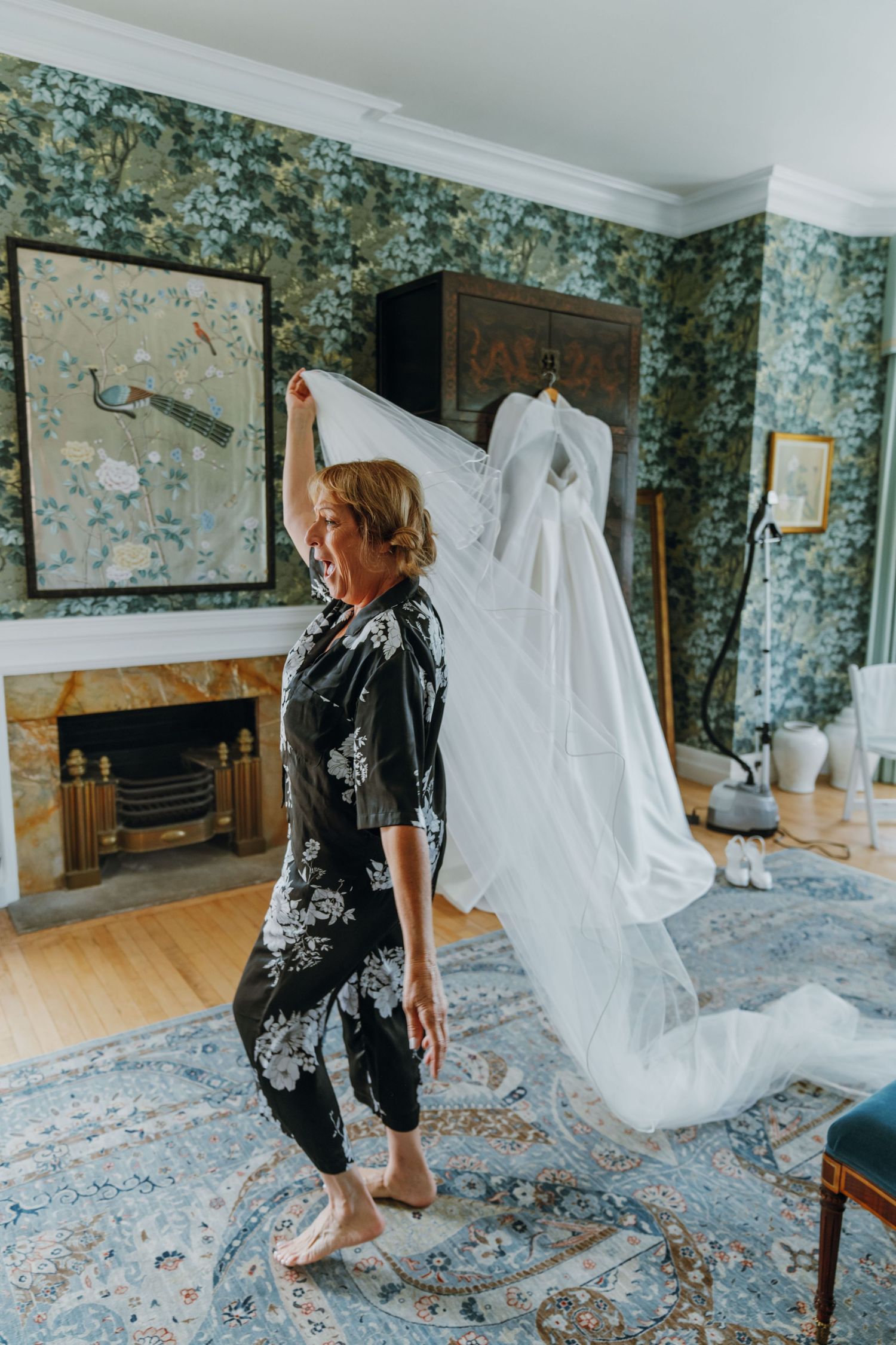 Woman in black floral pajamas holding up a white wedding dress veil in an elegant room with floral wallpaper. New Orleans, Louisiana, Missis
