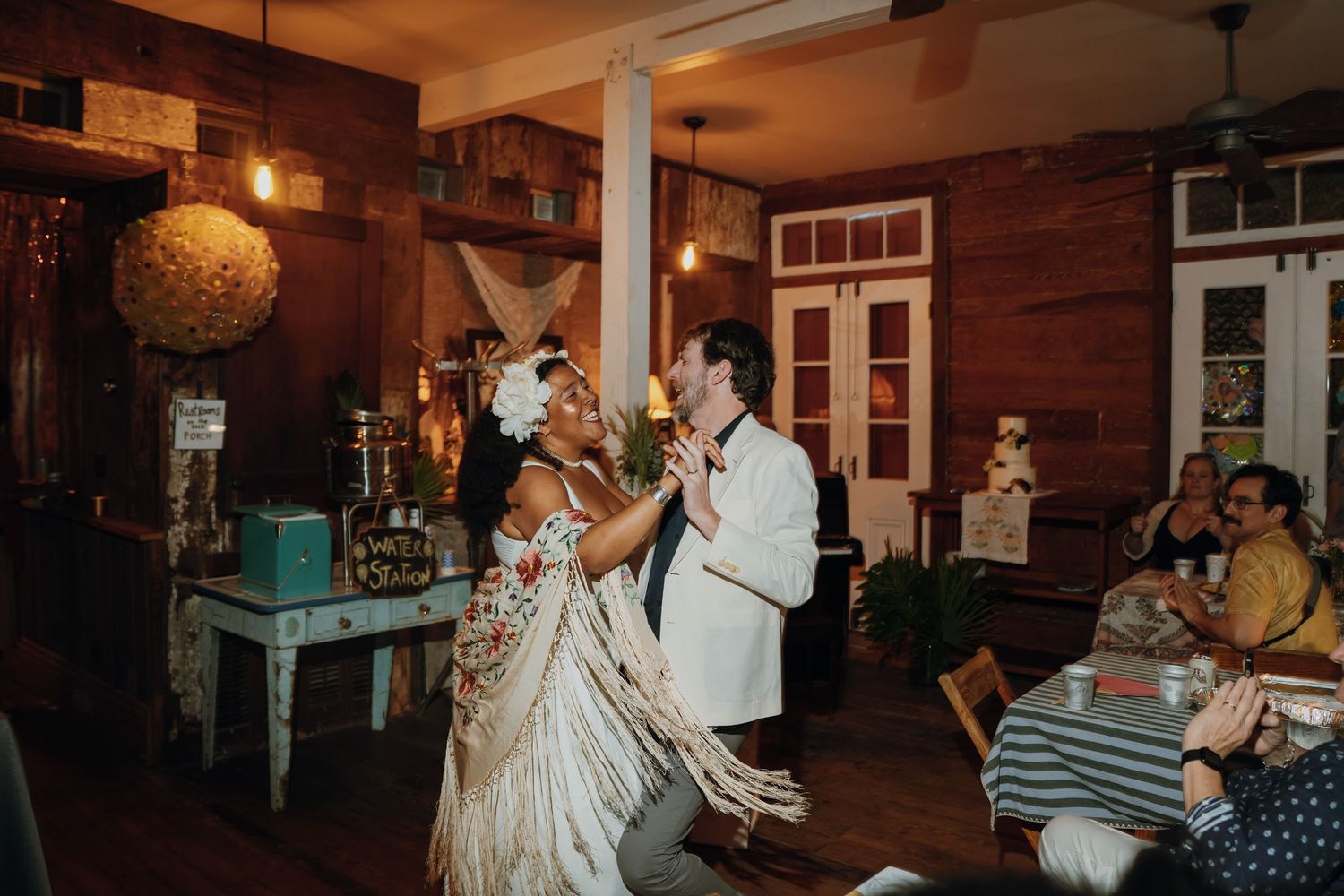 Bride in floral fringe shawl and groom in white jacket share first dance at rustic reception.