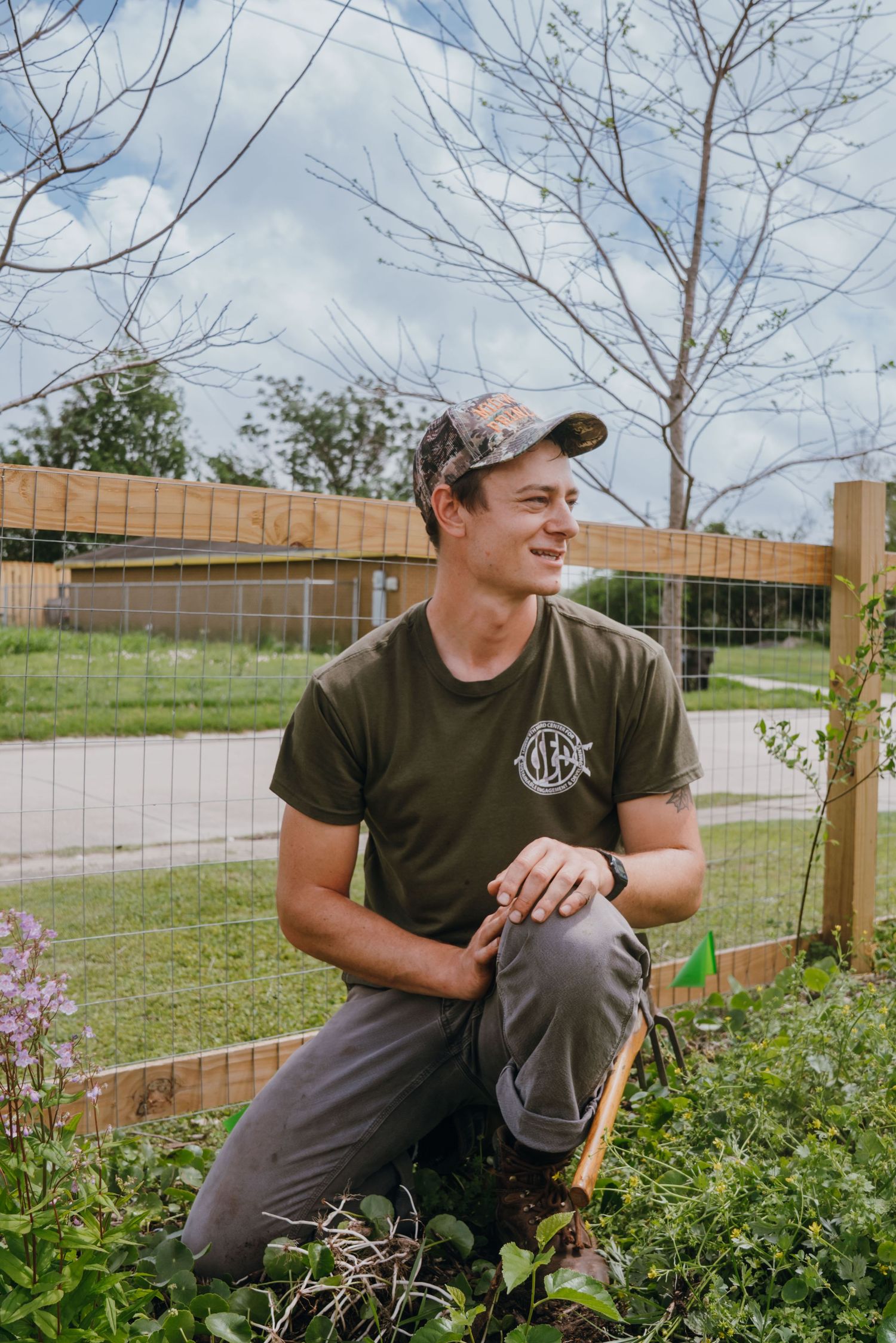 Smiling man in cap kneels in community garden with wooden fence.