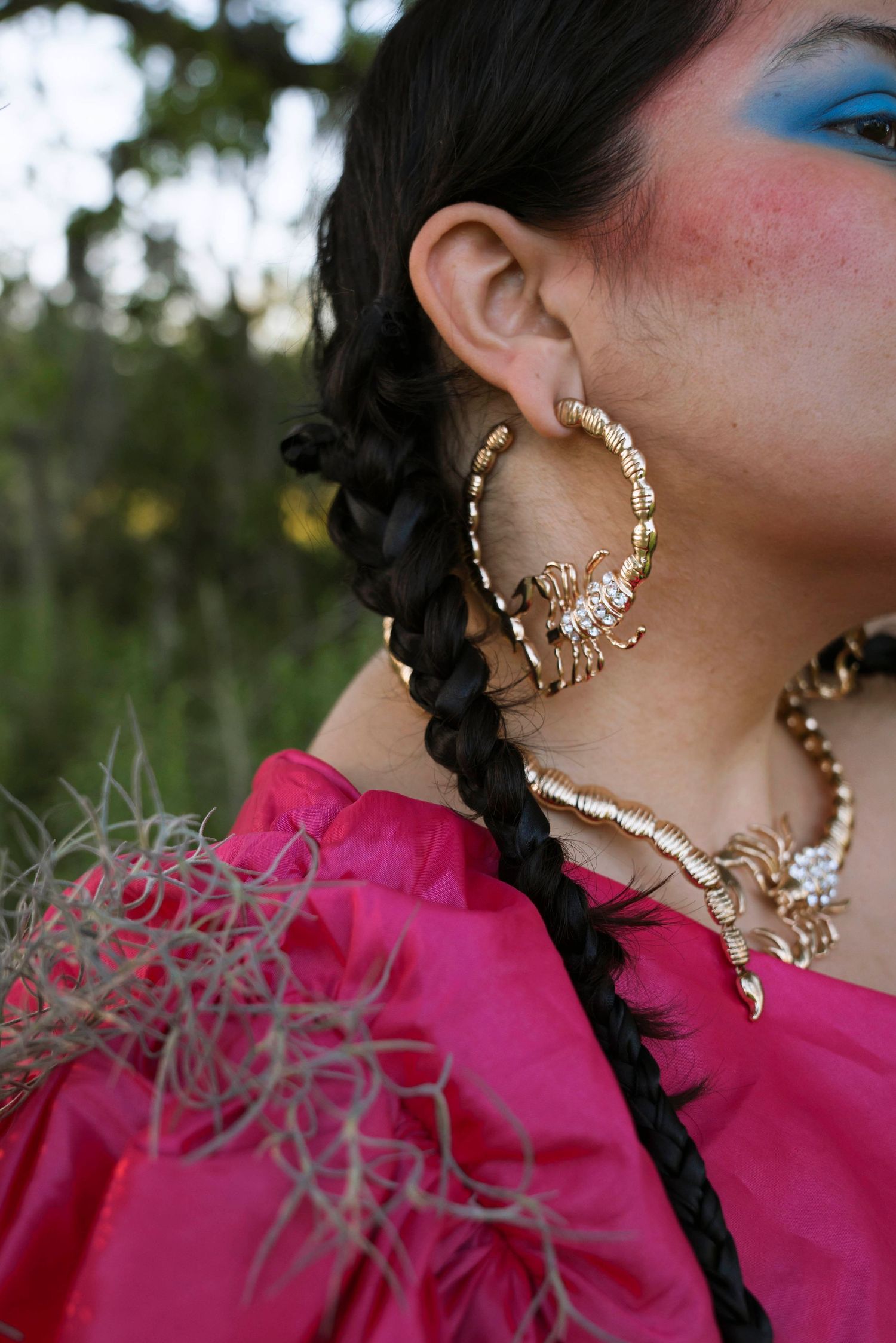 Close-up of gold scorpion hoop earring and necklace worn with blue eyeshadow and braided hair.