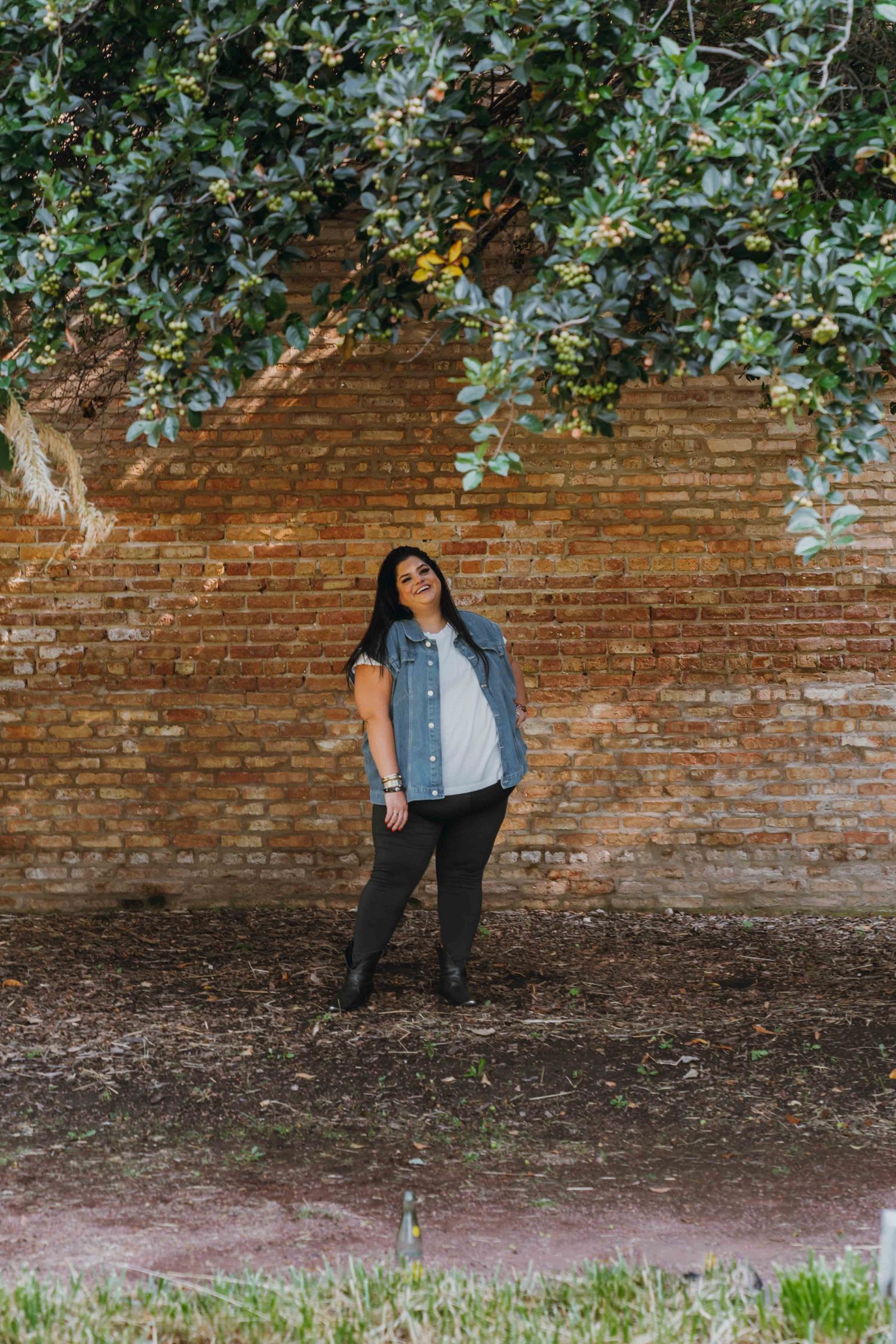 Woman in denim vest and black pants posing against a rustic brick wall with green foliage overhead.