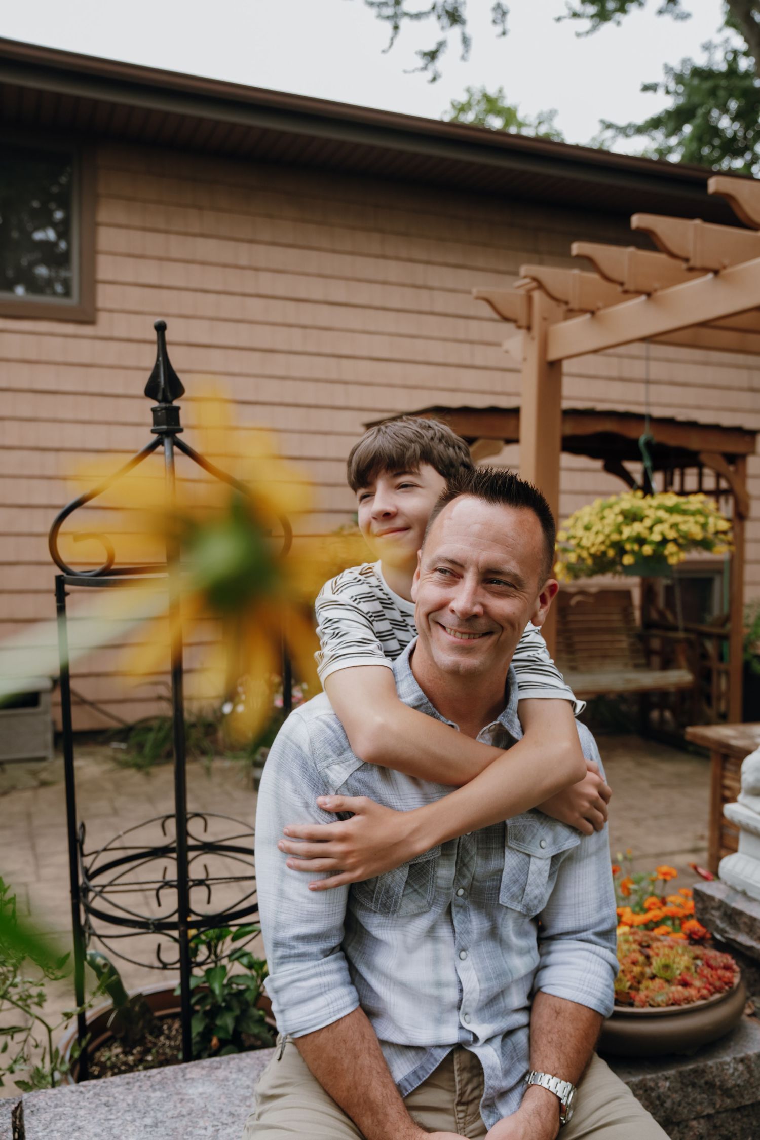 Smiling father and son hugging in backyard garden with sunflowers and pergola in background.