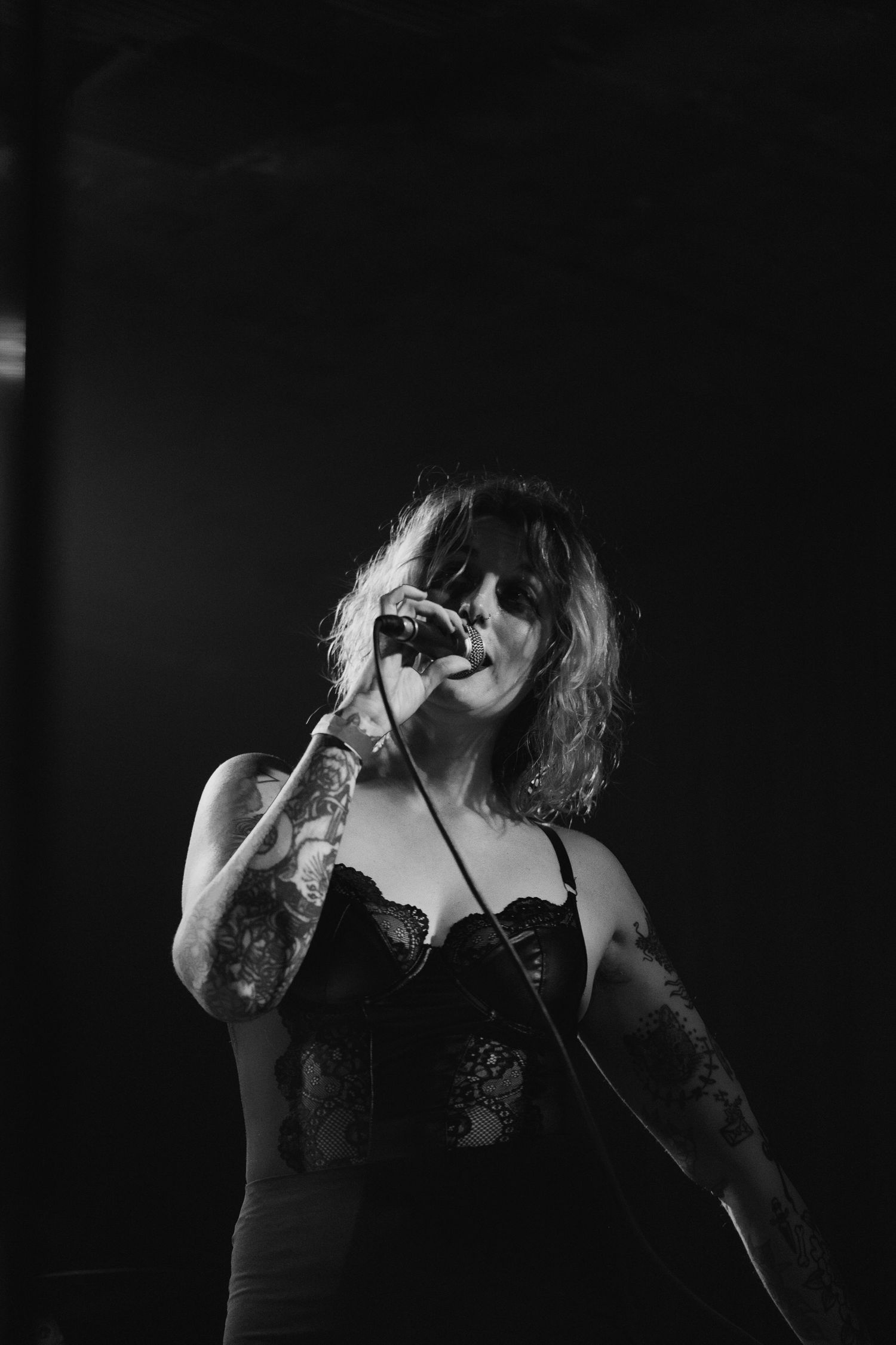 Black and white photo of tattooed female singer performing on dark stage with microphone.
