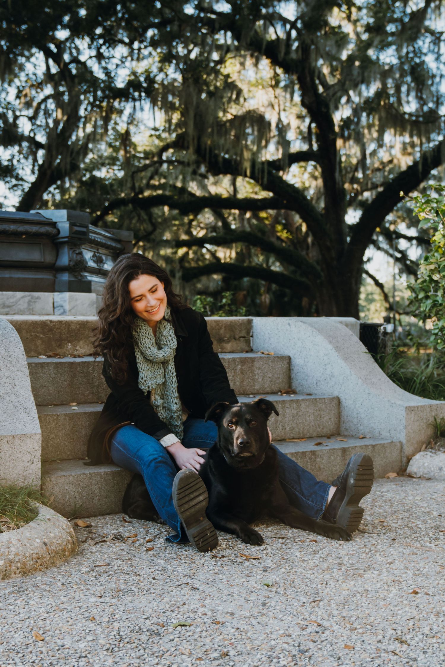 A smiling woman sits on stone steps with a black Labrador dog, surrounded by Spanish moss-draped trees in a park.
