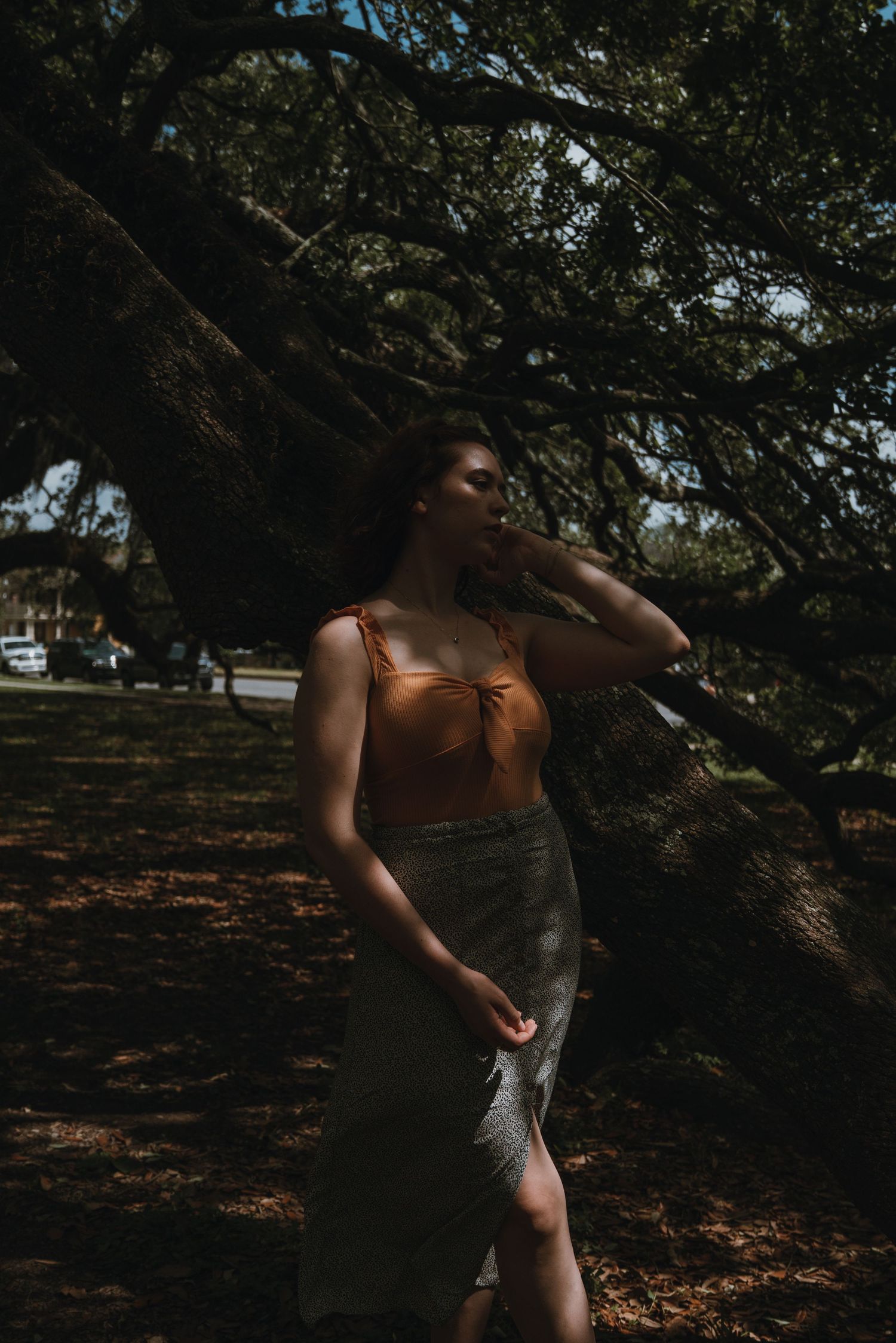 Woman in orange top and gray skirt standing under dramatic tree branches in dappled sunlight.