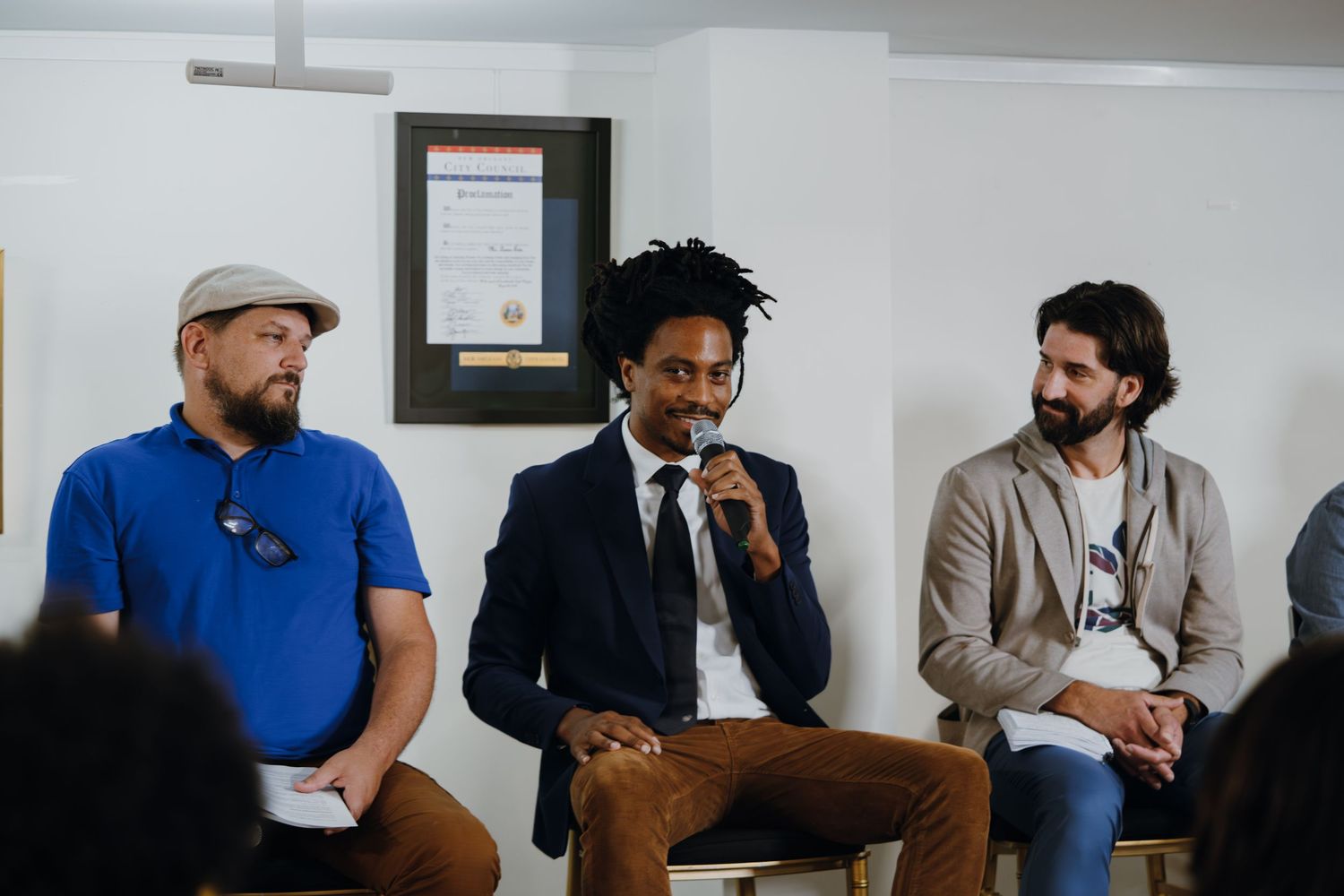 Three men seated in a panel discussion, with a man with dreadlocks speaking into a microphone at a community event.