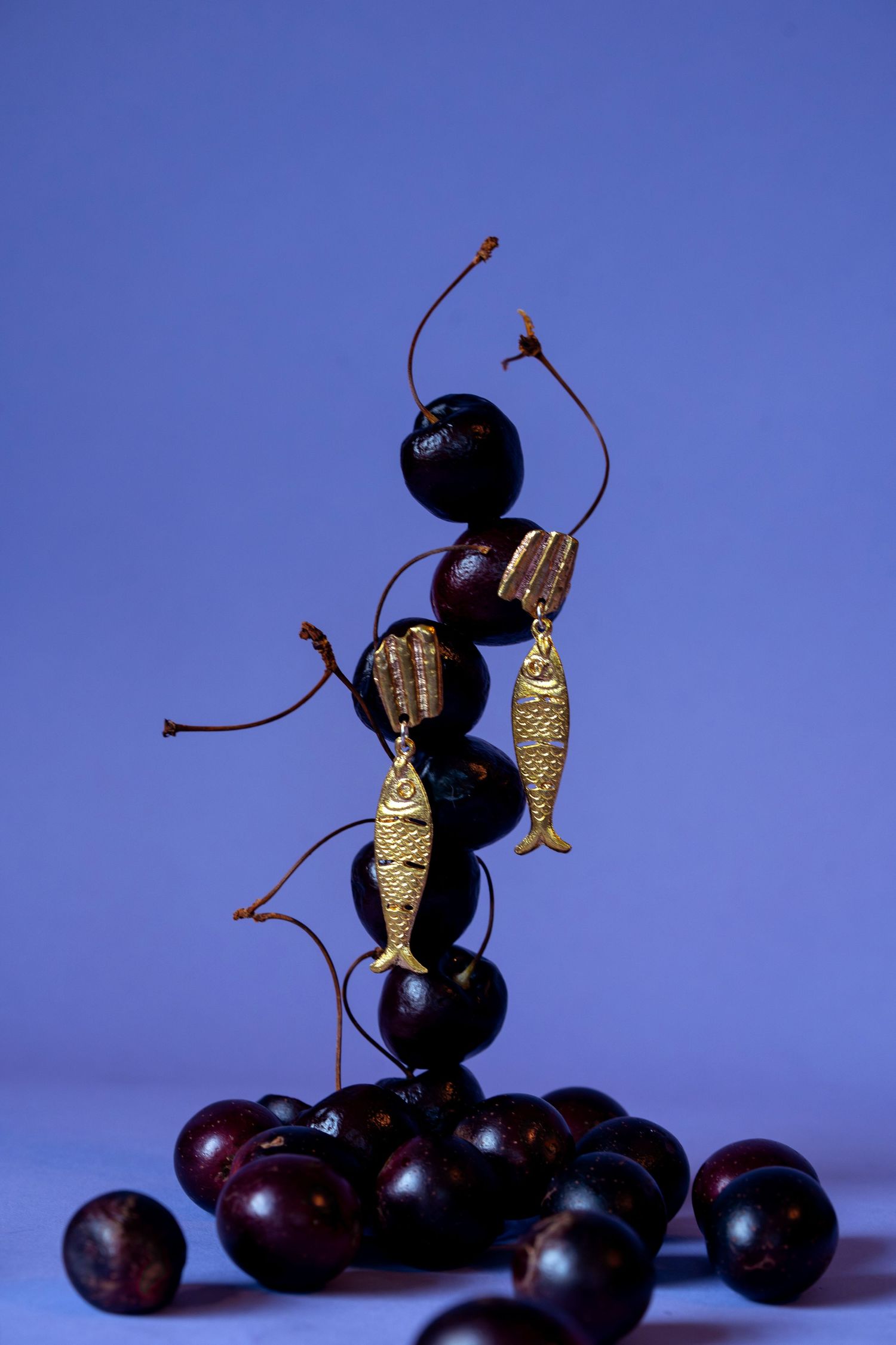 Gold fish charm earrings balanced on a stacked tower of dark cherries on blue background.