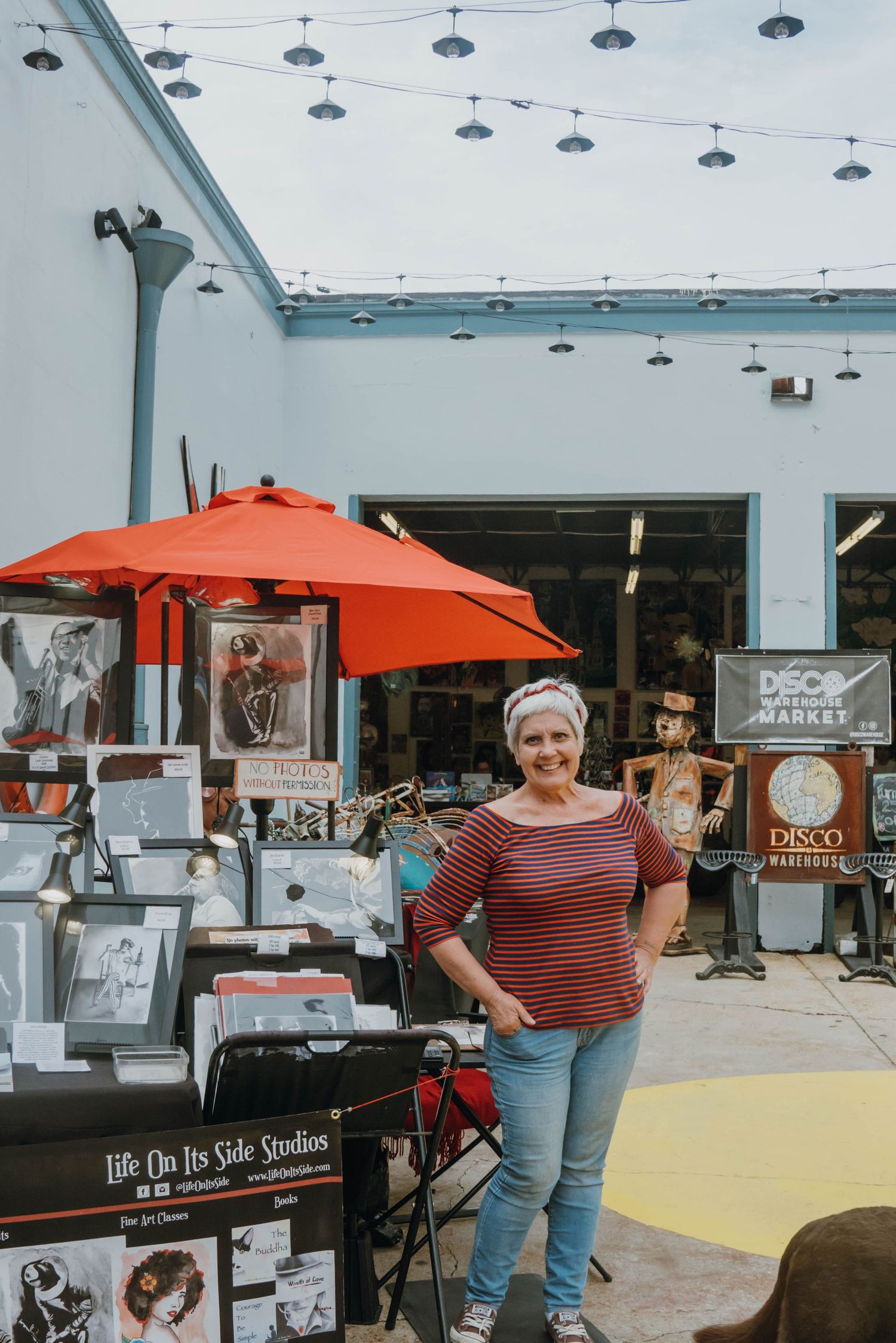 Woman in striped shirt standing outside Deco Warehouse Market with art prints and vintage items displayed.
