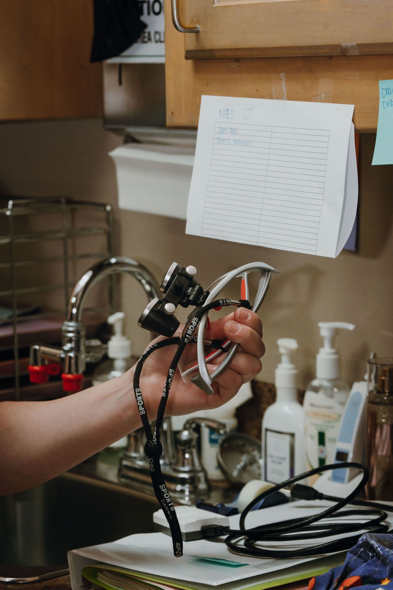 A hand holds dental loupes with a lanyard near a sink in a dental office workspace.
