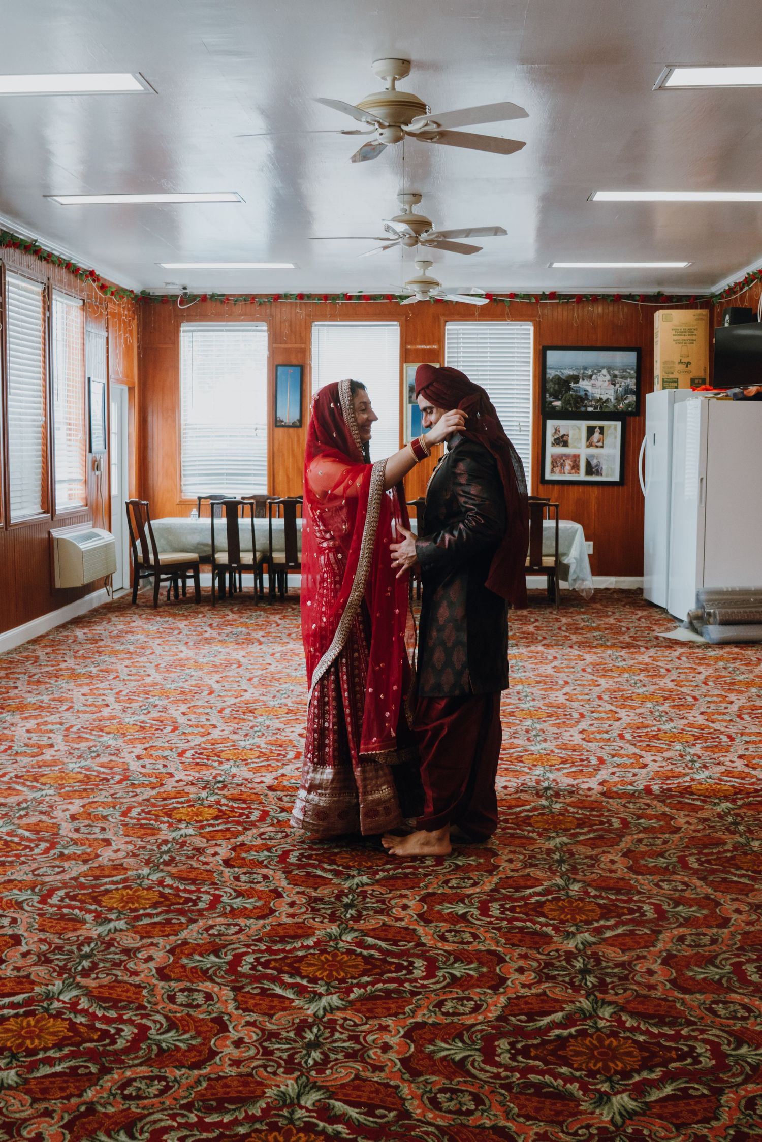 Indian bride and groom in traditional wedding attire facing each other on vibrant red patterned carpet.
