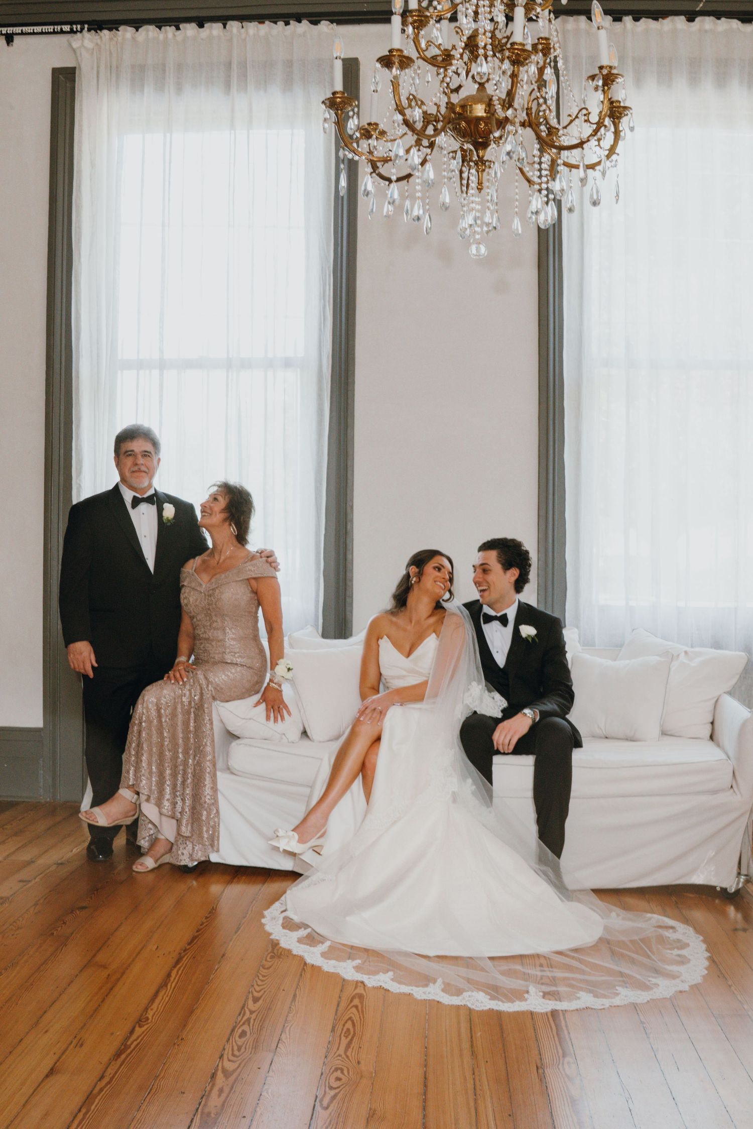 Bride and groom posing with parents on white sofa under crystal chandelier in bright elegant wedding room.