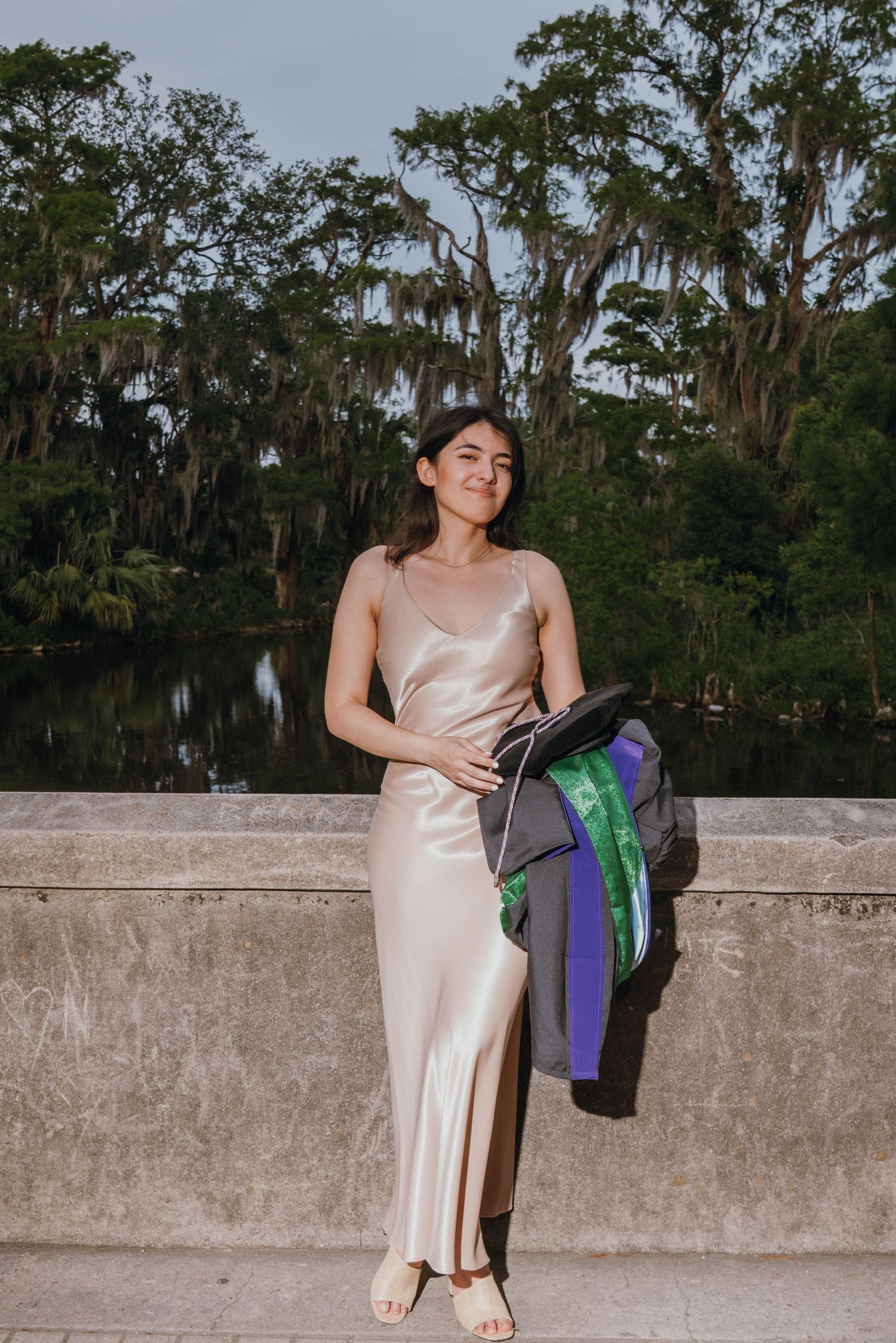Young woman in satin slip dress holding graduation gown beside scenic tree-lined waterway.