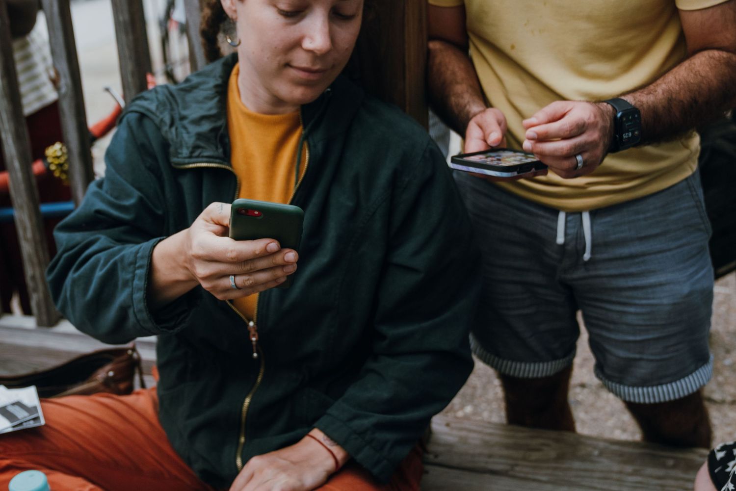 Young woman in green jacket looking at smartphone while man in yellow shirt checks his phone nearby.