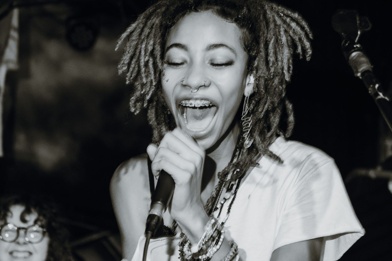 Black and white photo of a woman with dreadlocks singing passionately into a microphone on stage.