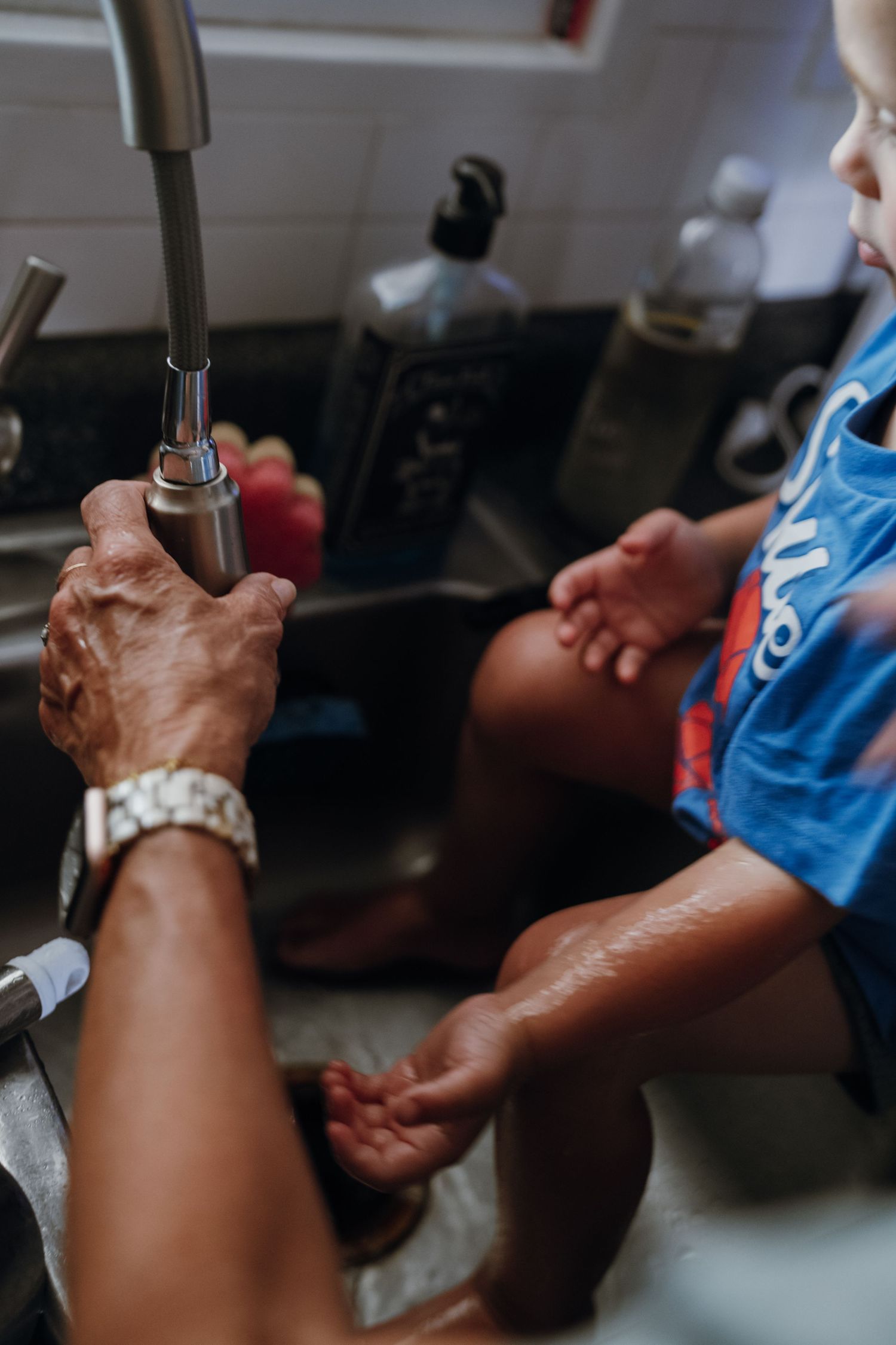 Adult washing a young child's hands at a kitchen sink with running water.