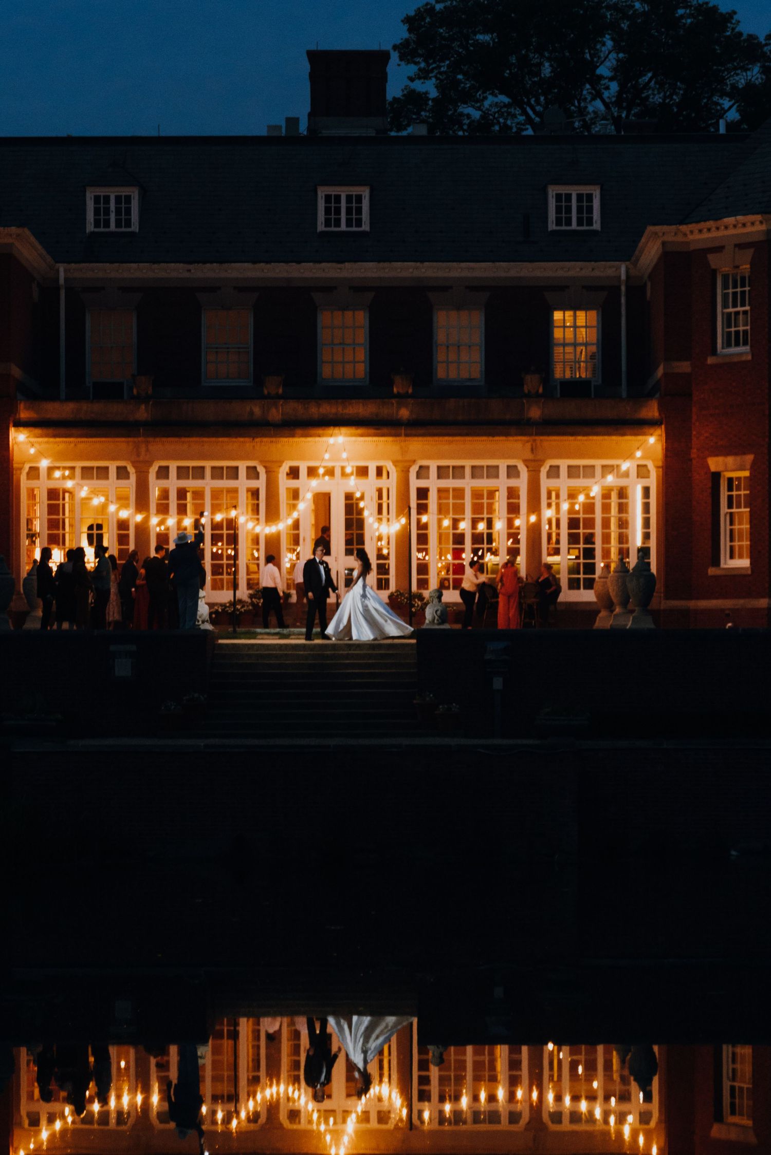 Bride and groom stand illuminated by warm string lights outside a grand estate reflected in a pond at night. New Orleans, Louisiana, Mississ