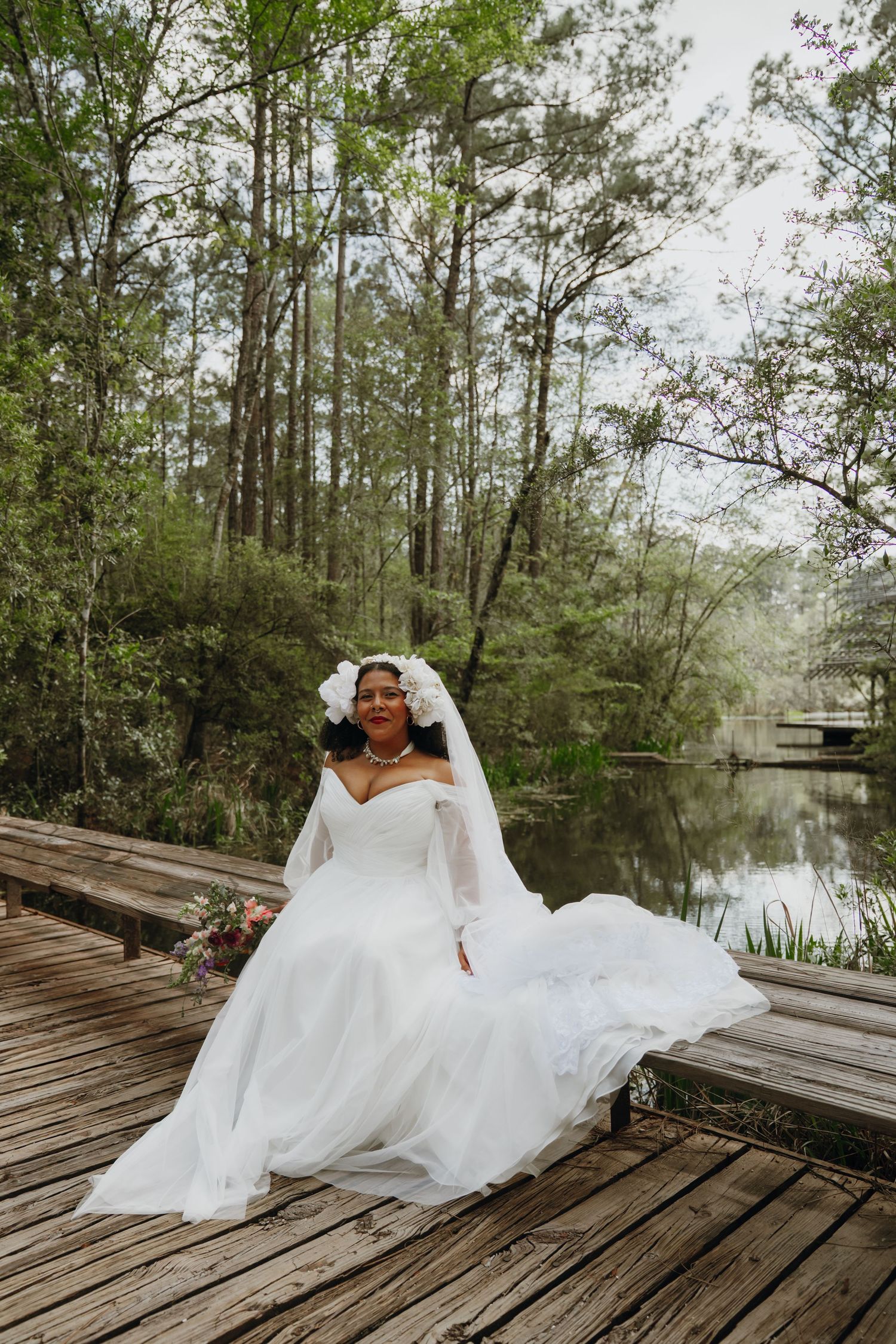 Radiant bride in white gown and floral headpiece sits on wooden lakeside dock.
