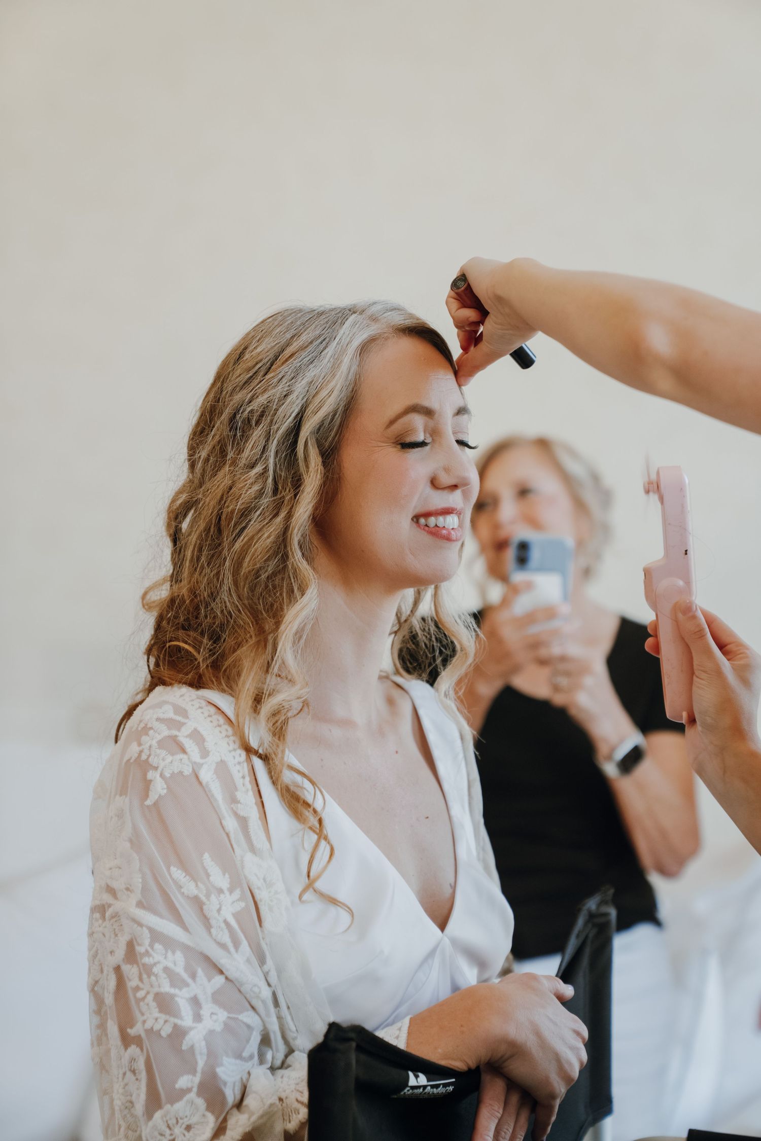 Smiling bride getting makeup applied while bridesmaid photographs the moment in background.