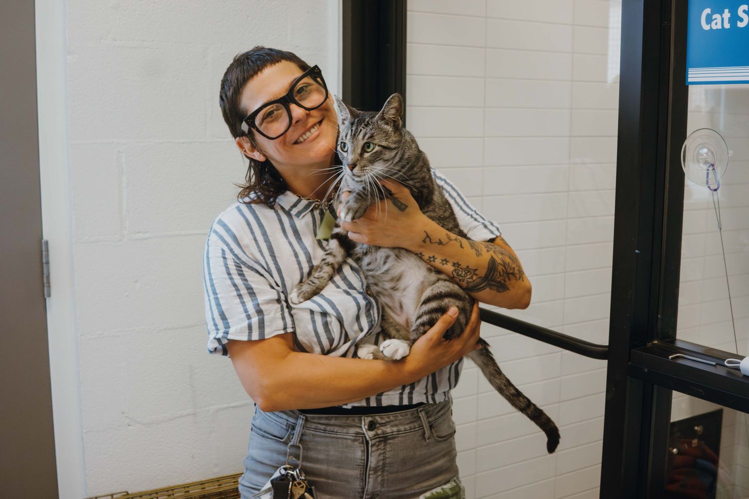 A smiling woman with glasses and tattoos holds a gray tabby cat in a white-tiled room near a glass door.