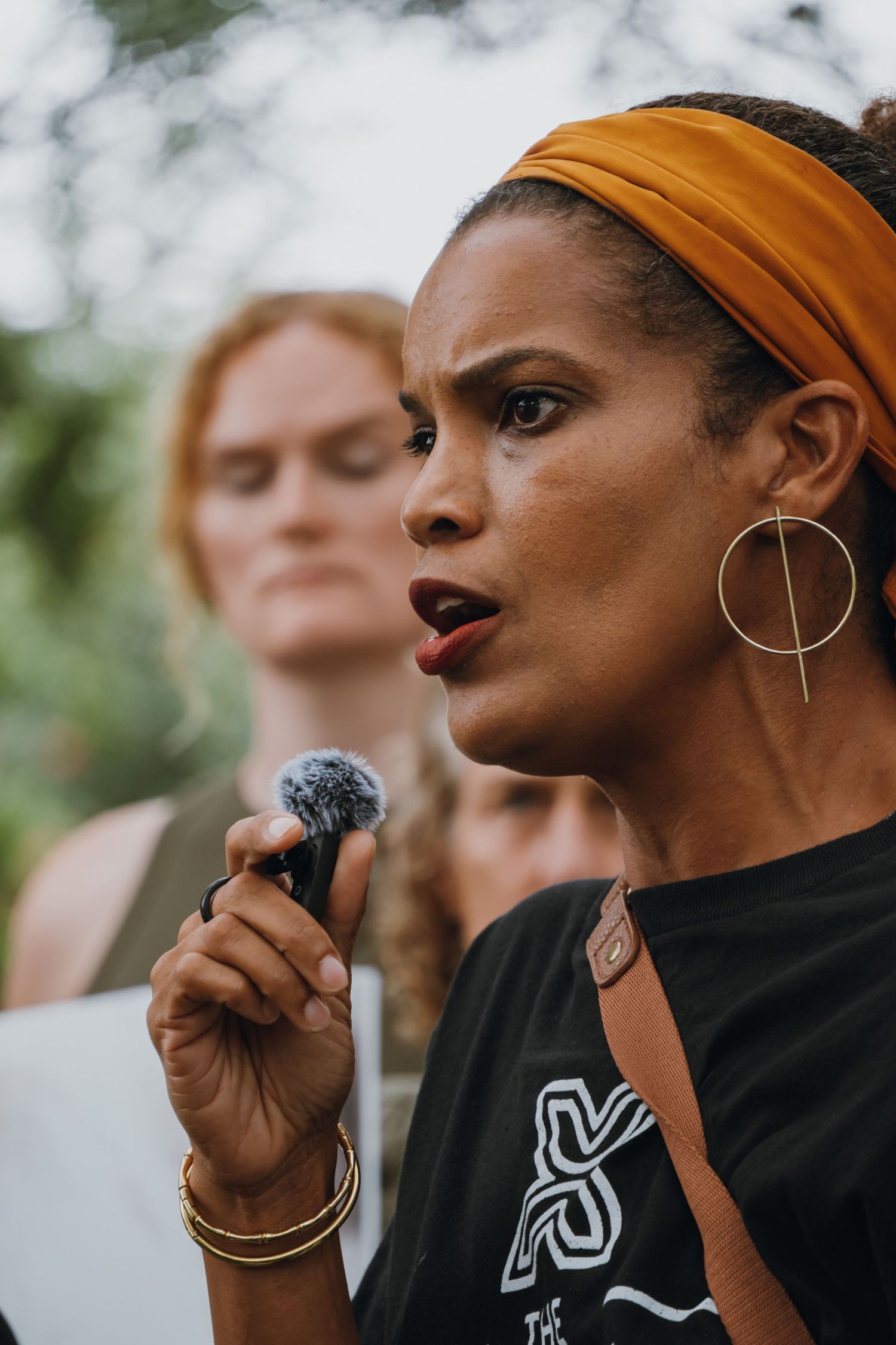 Woman wearing an orange headband speaking passionately into a microphone at an outdoor gathering.