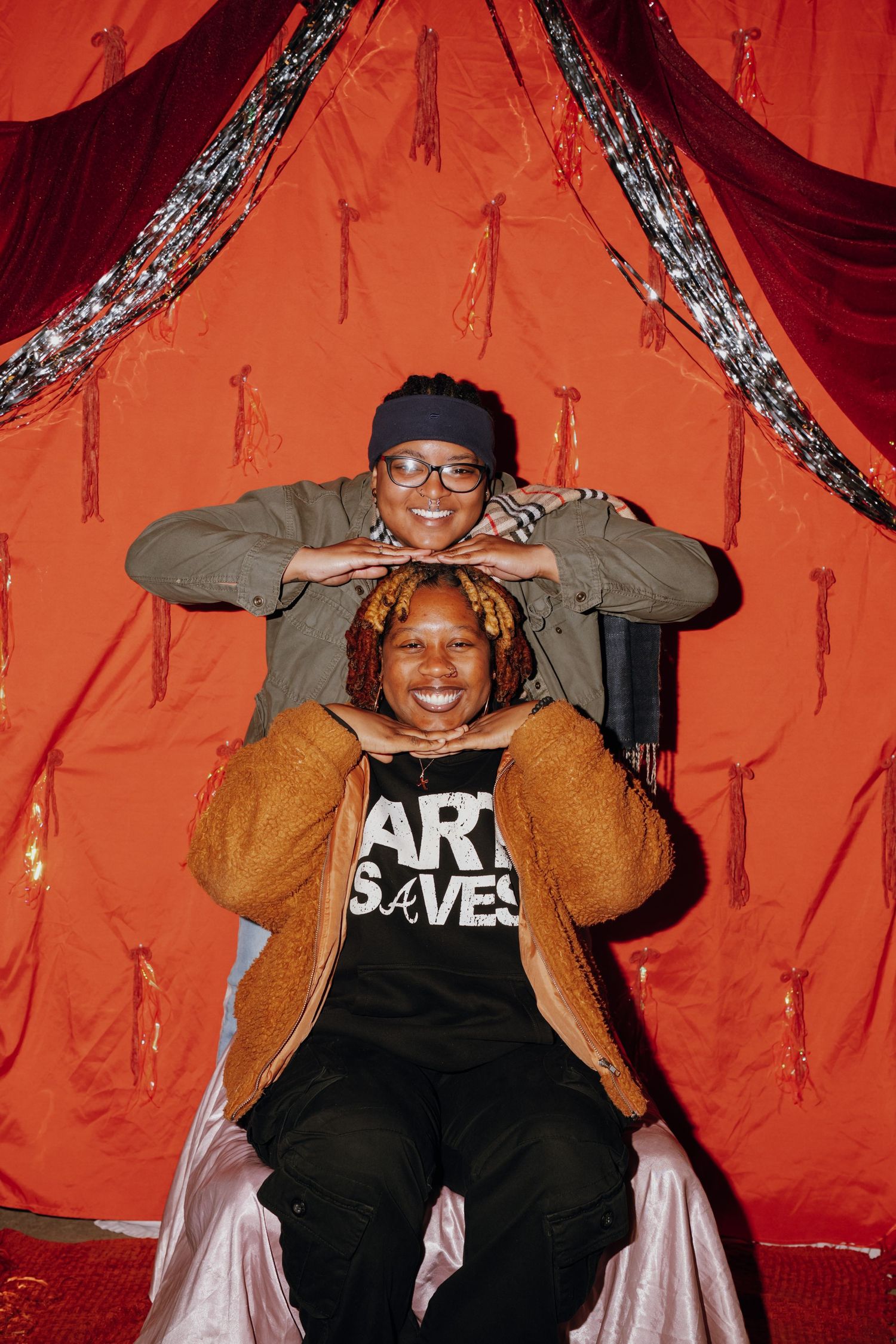 Two friends pose playfully in front of a red draped backdrop, one wearing an Art Saves shirt.