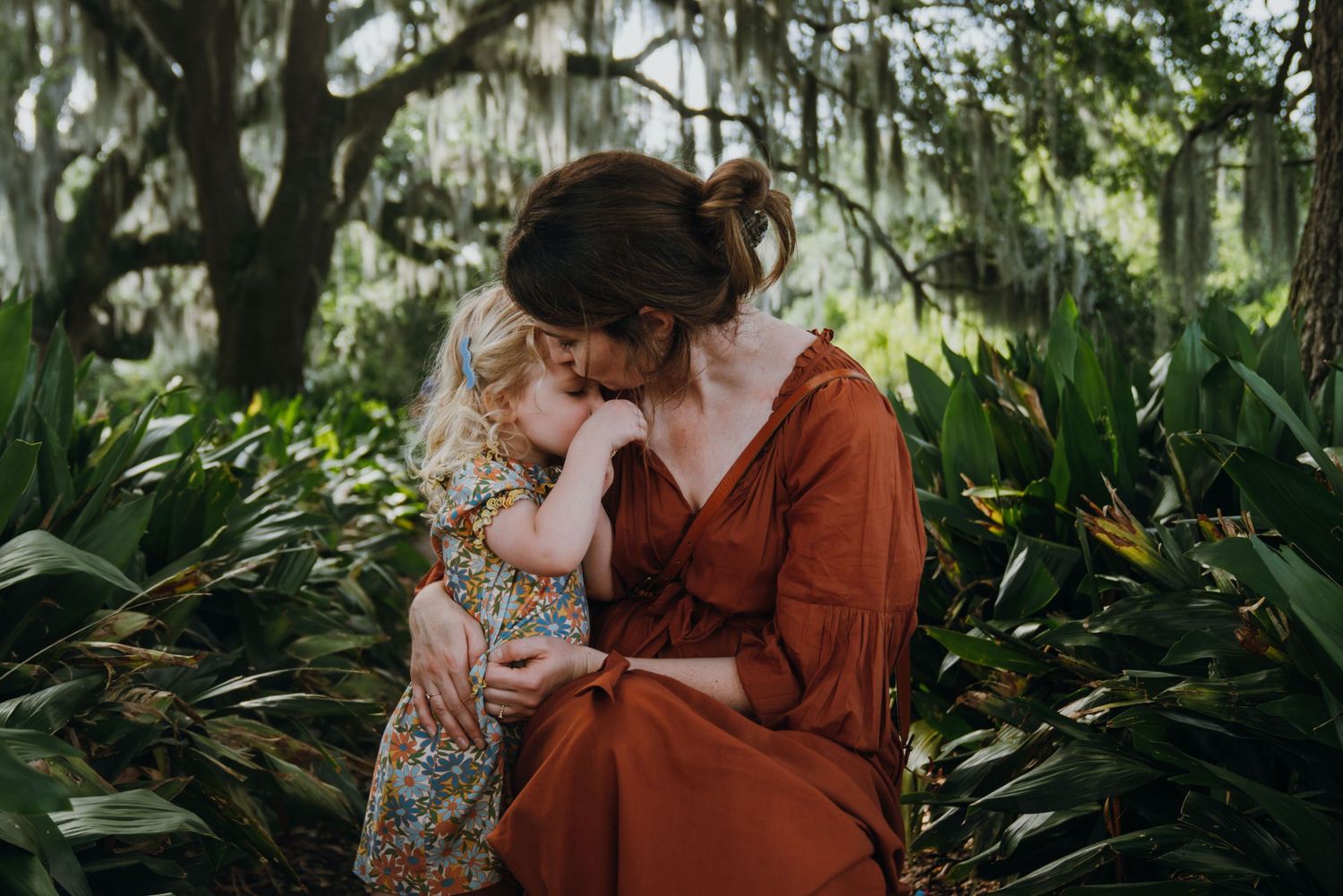 A mother in a rust dress tenderly embraces her young daughter outdoors among lush green foliage.