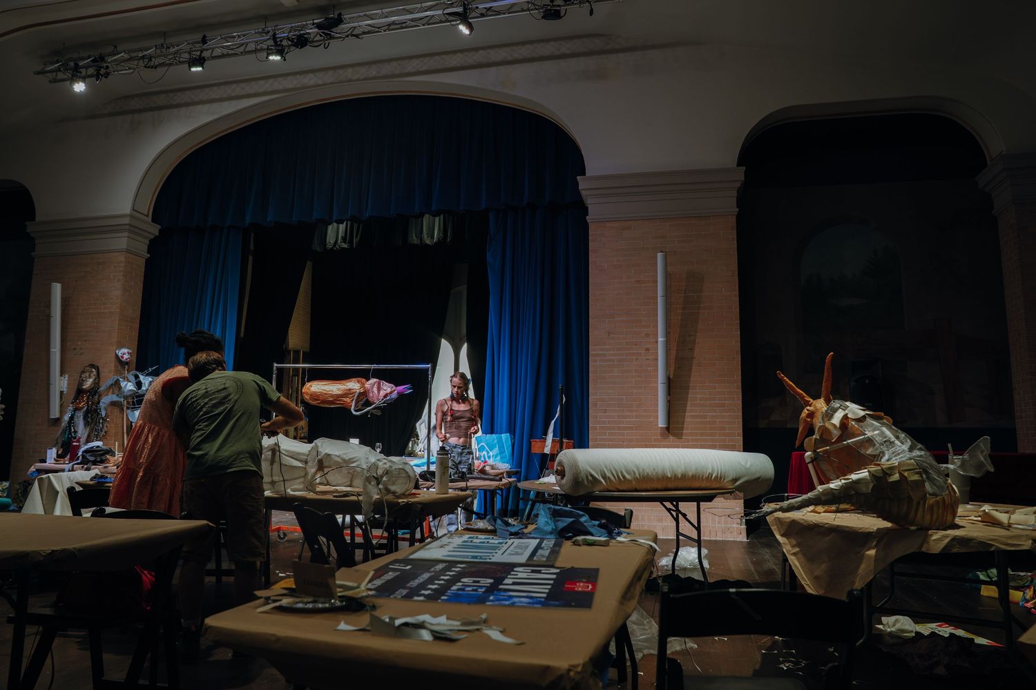 Backstage theater scene with blue curtains, costumes, and props on tables in a dimly lit performance hall.
