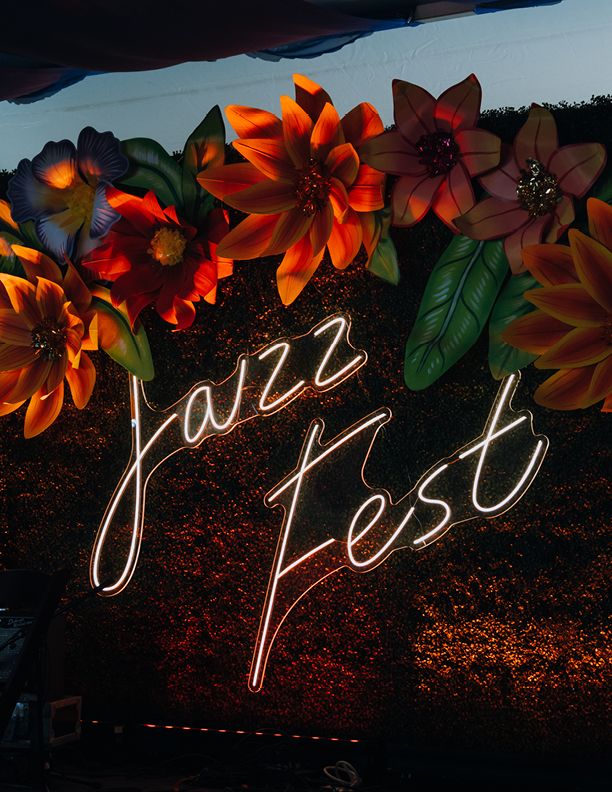 Glowing neon Jazz Fest sign surrounded by vibrant large artificial flowers in orange, red, and pink hues at a festival.