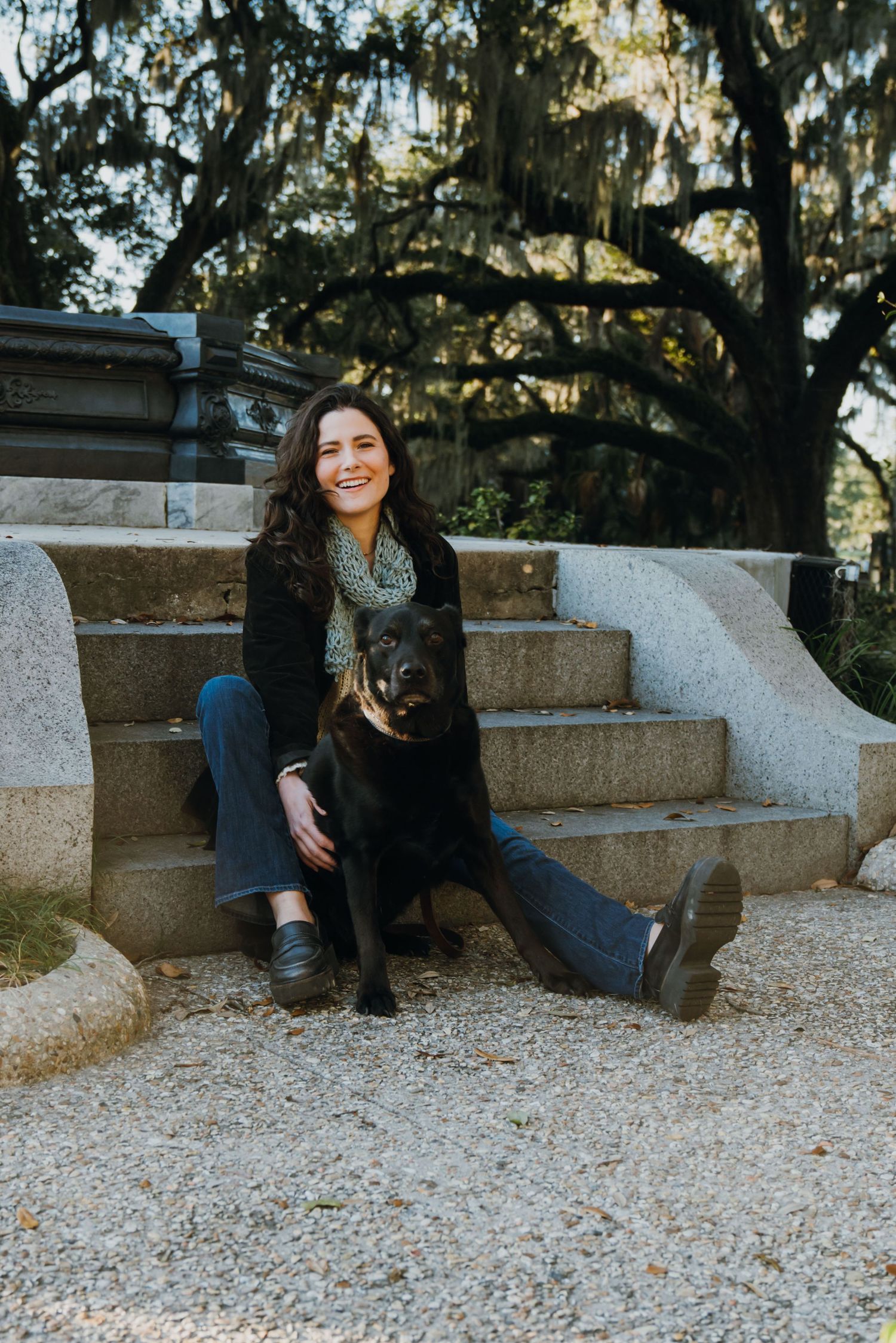 Smiling woman sitting on stone steps with a black dog, surrounded by moss-draped trees in a scenic outdoor setting.