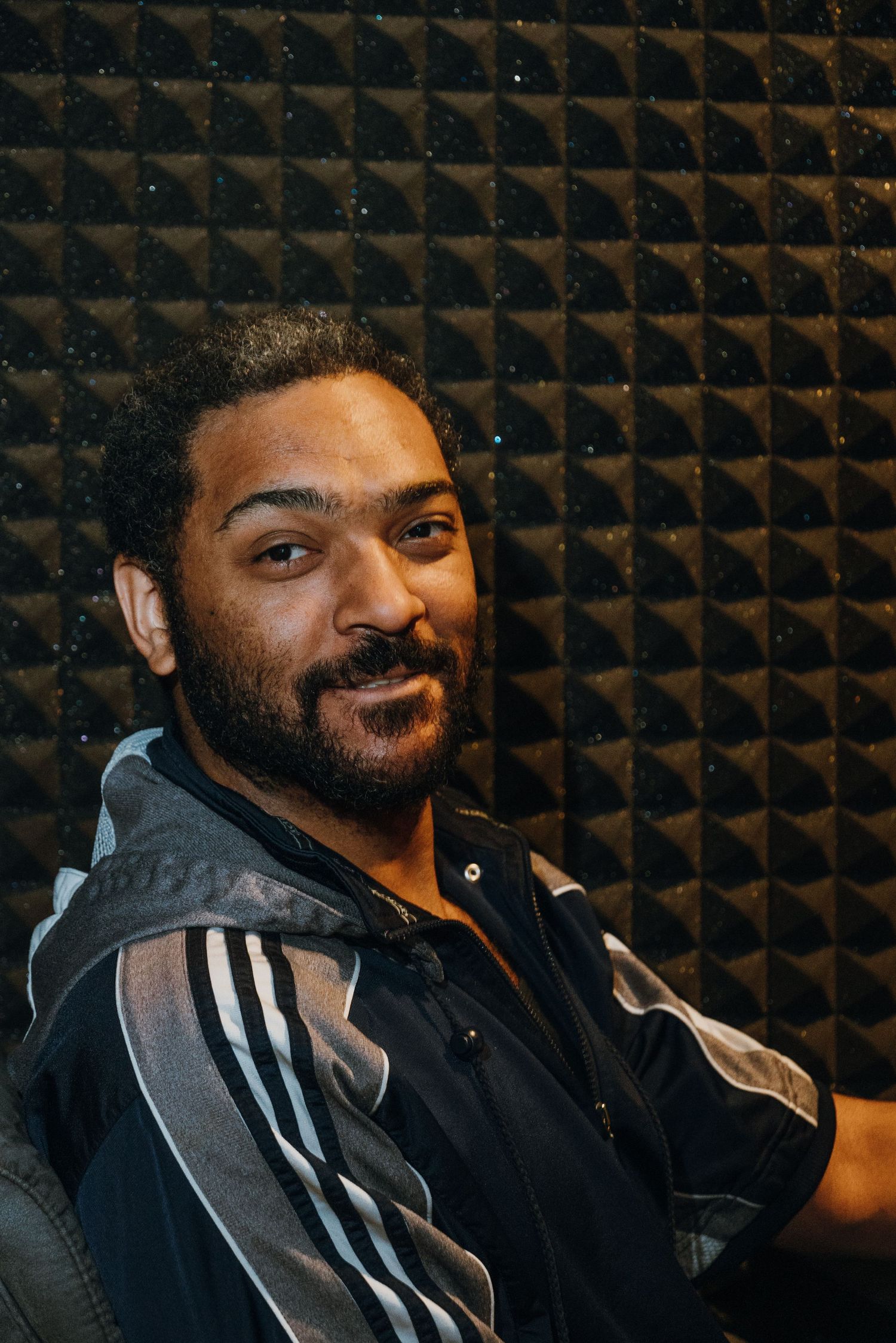 Man with beard smiling in front of dark acoustic foam wall in a professional recording studio.