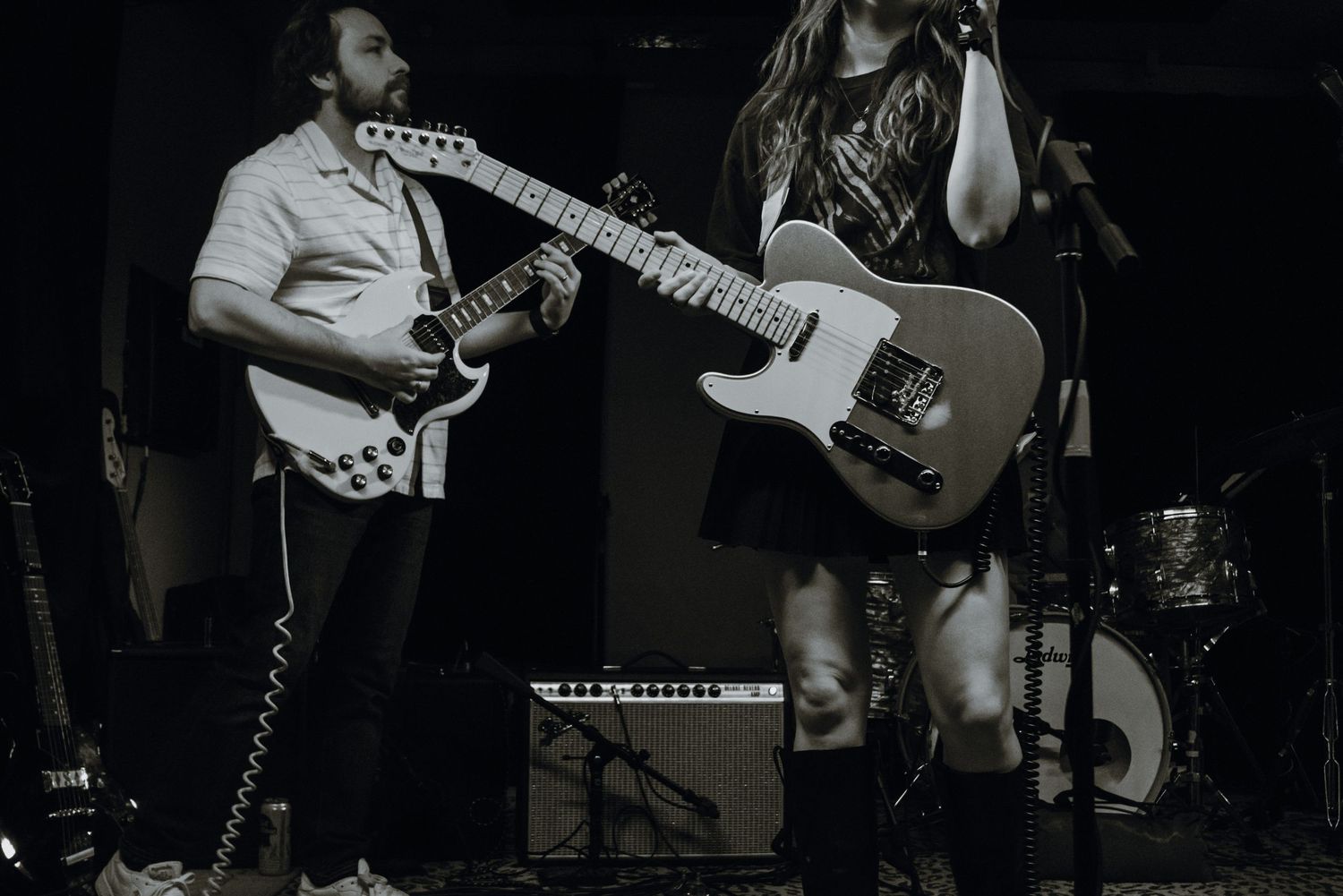Black and white photo of two guitarists performing energetically on stage under dim lighting.