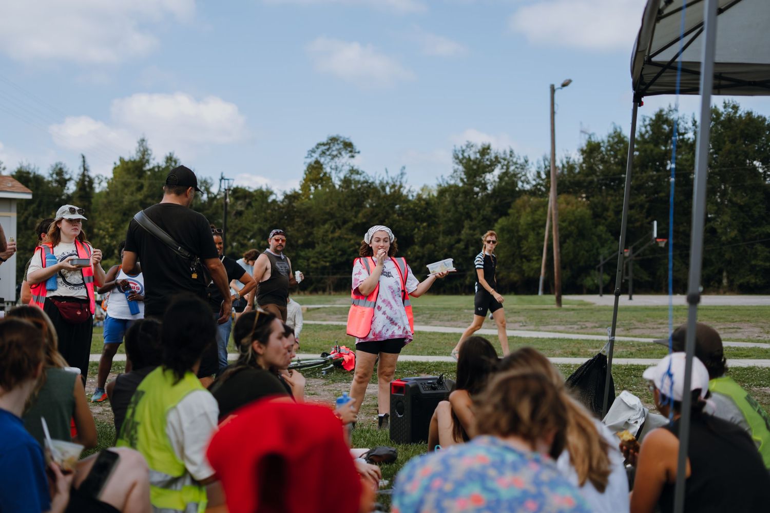 Woman in pink wig speaks to a crowd gathered outdoors at a community cycling event.