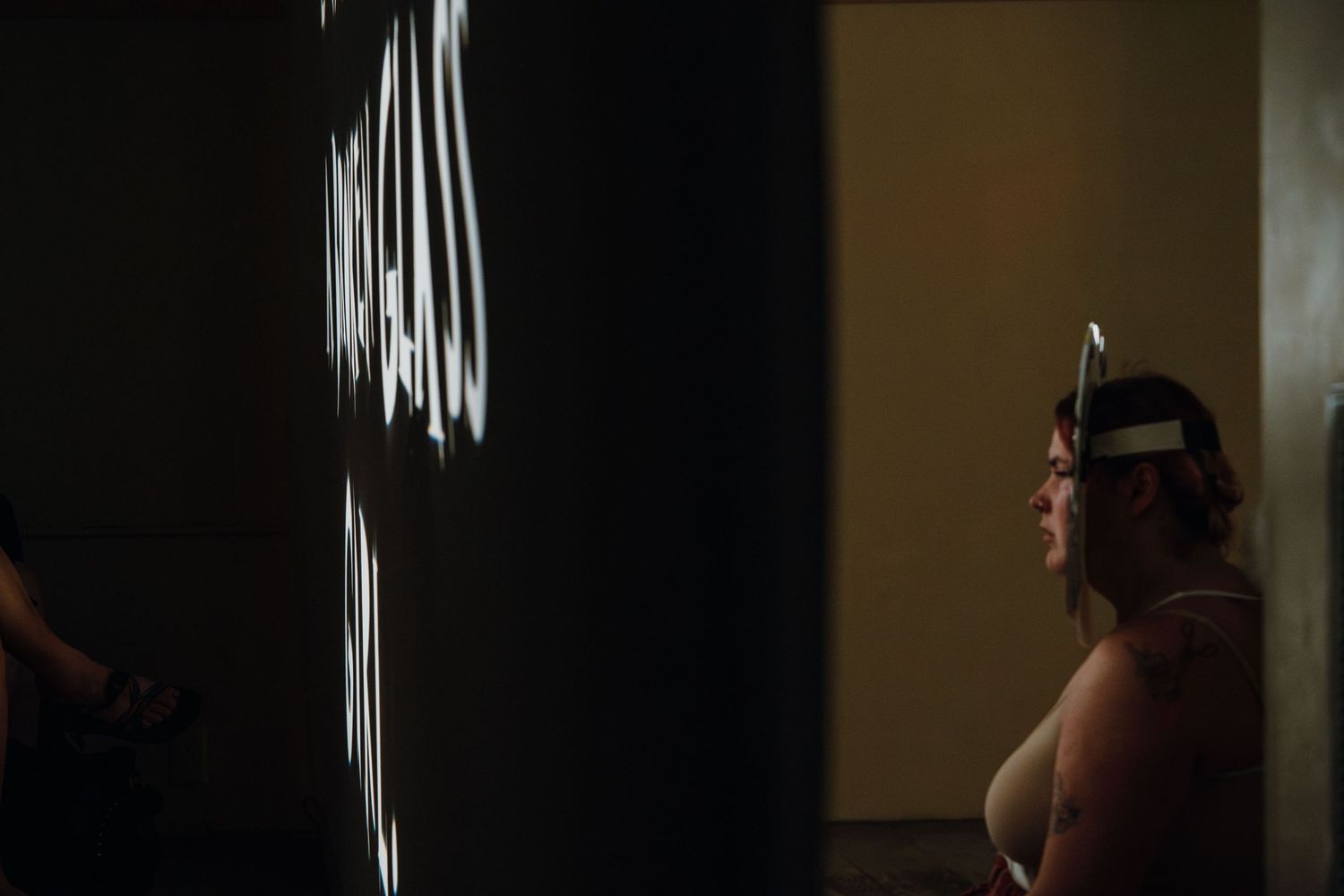 Woman with headband gazes at projected white text on dark wall in performance.