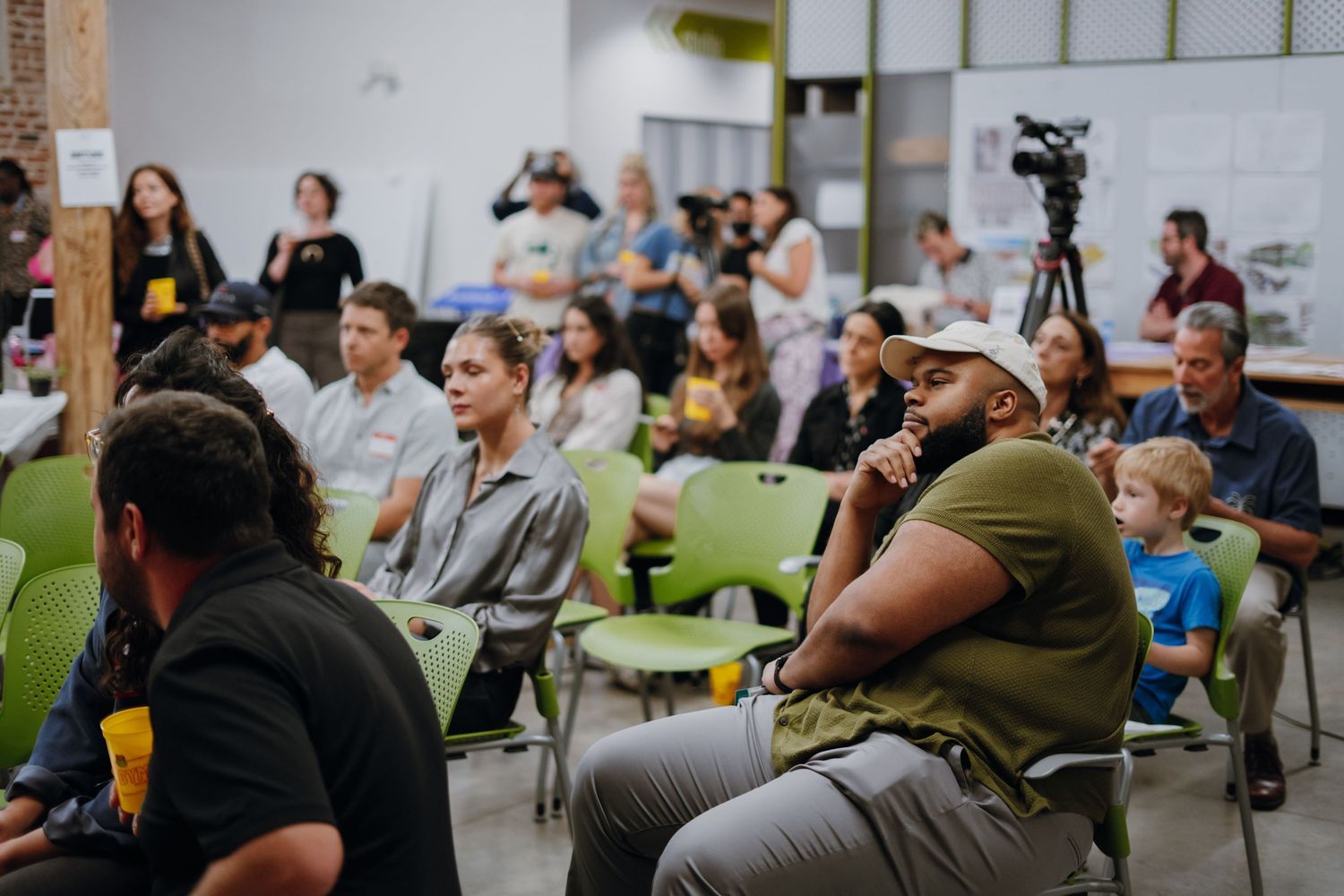 Diverse audience seated in green chairs attentively watching a community presentation indoors.