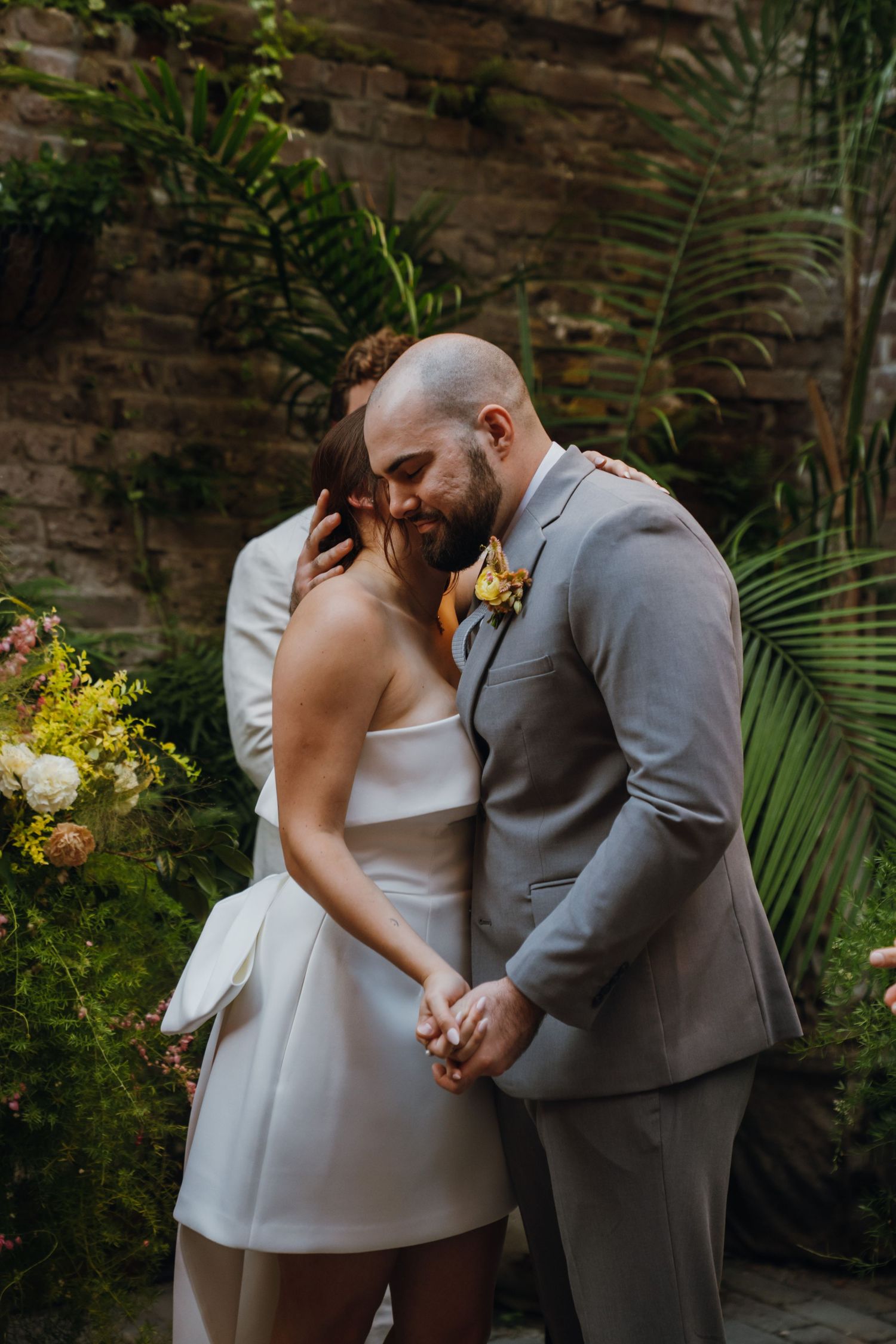 Newlyweds embrace tenderly during outdoor wedding ceremony with tropical palm backdrop and floral arrangements. New Orleans, Louisiana, Miss