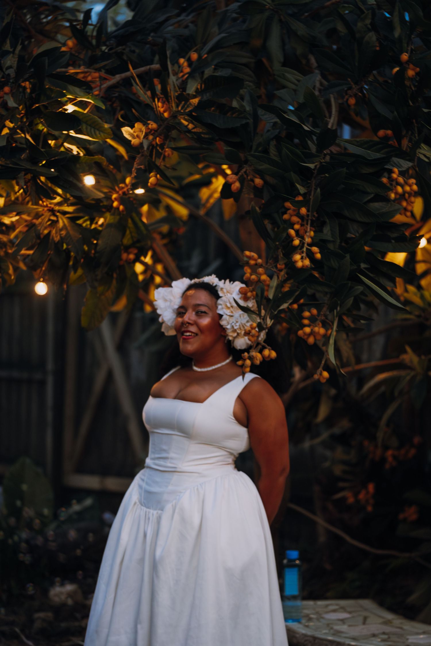Smiling bride in white dress and floral crown stands under fruit tree with string lights at night.