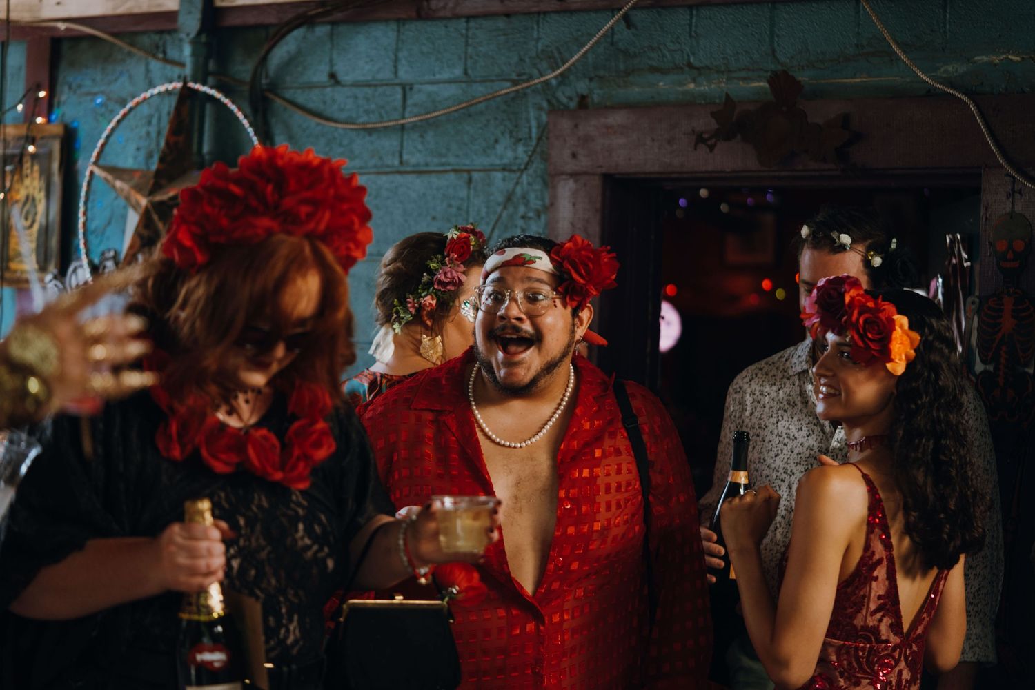 Group of guests in vibrant red costumes and floral headpieces laugh and celebrate at a lively party.