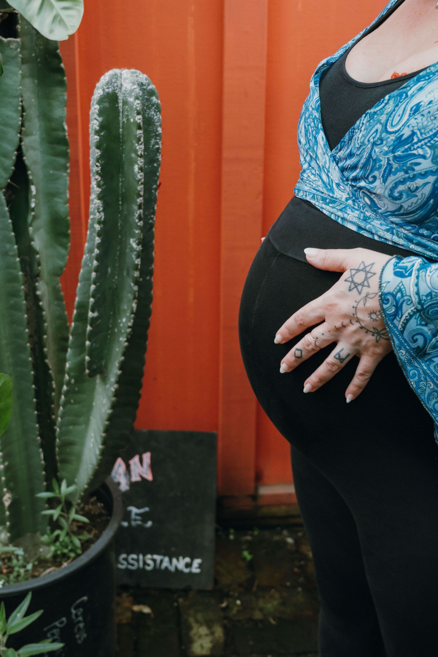 Close-up of pregnant belly with tattooed hand resting on bump beside tall cactus outdoors.